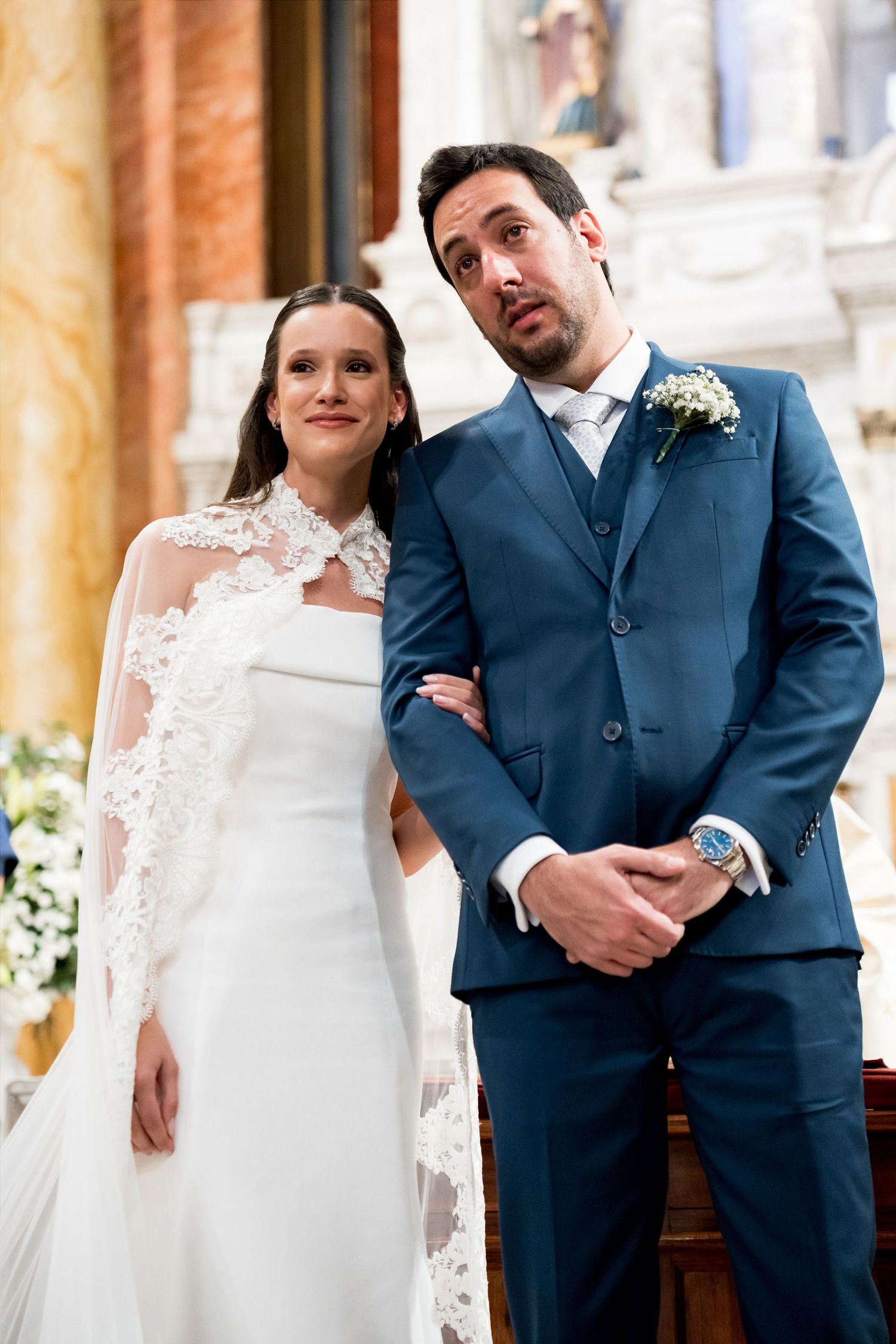 Bride in white lace gown and groom in blue suit standing together at church wedding ceremony altar.