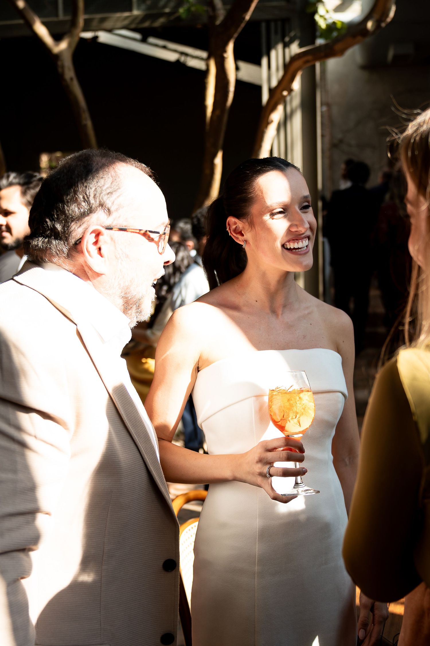 Woman in white strapless dress laughing and holding a wine glass at an outdoor social event.
