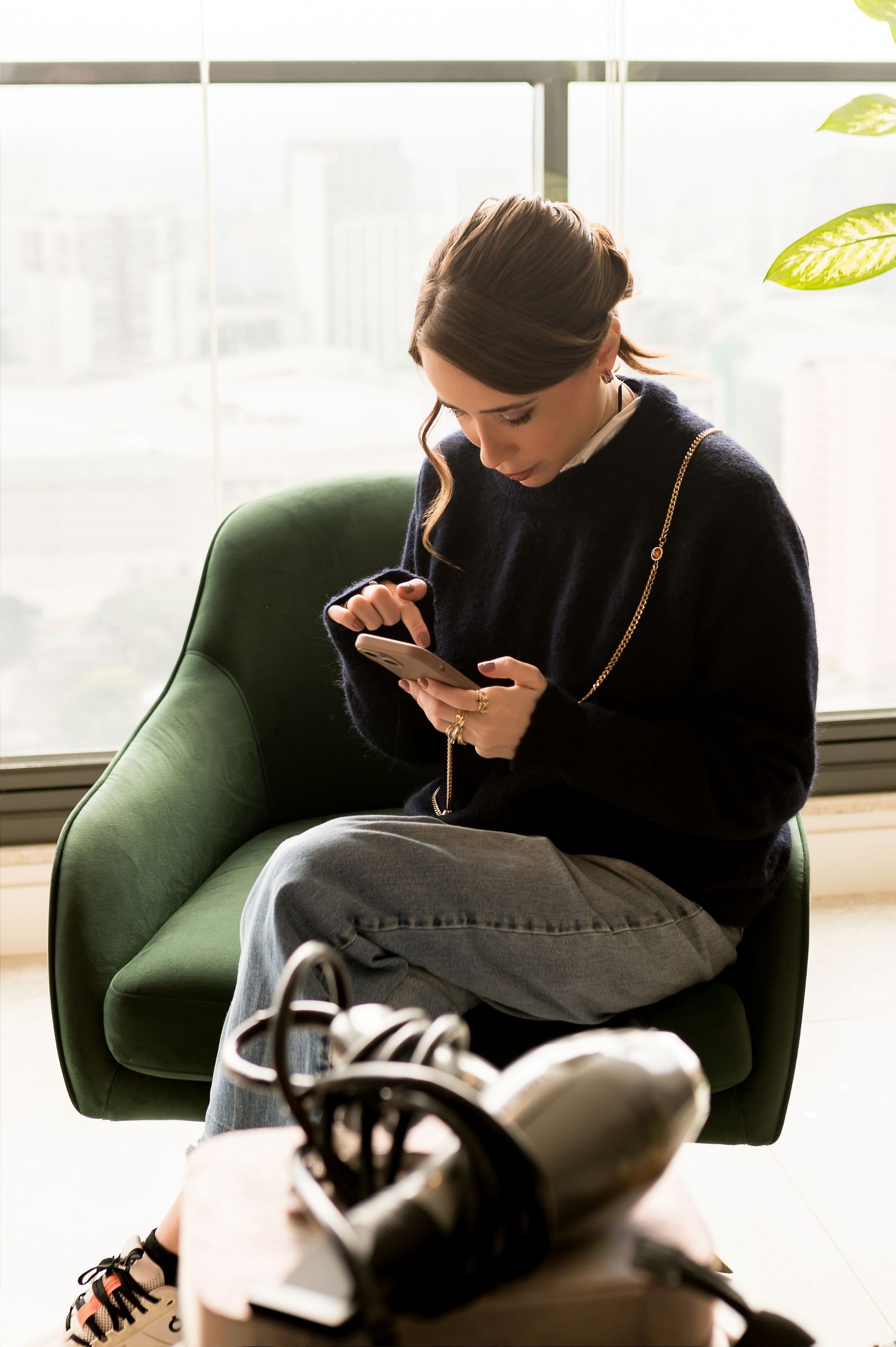 Young woman in navy sweater and jeans sitting in green armchair using smartphone near bright window.