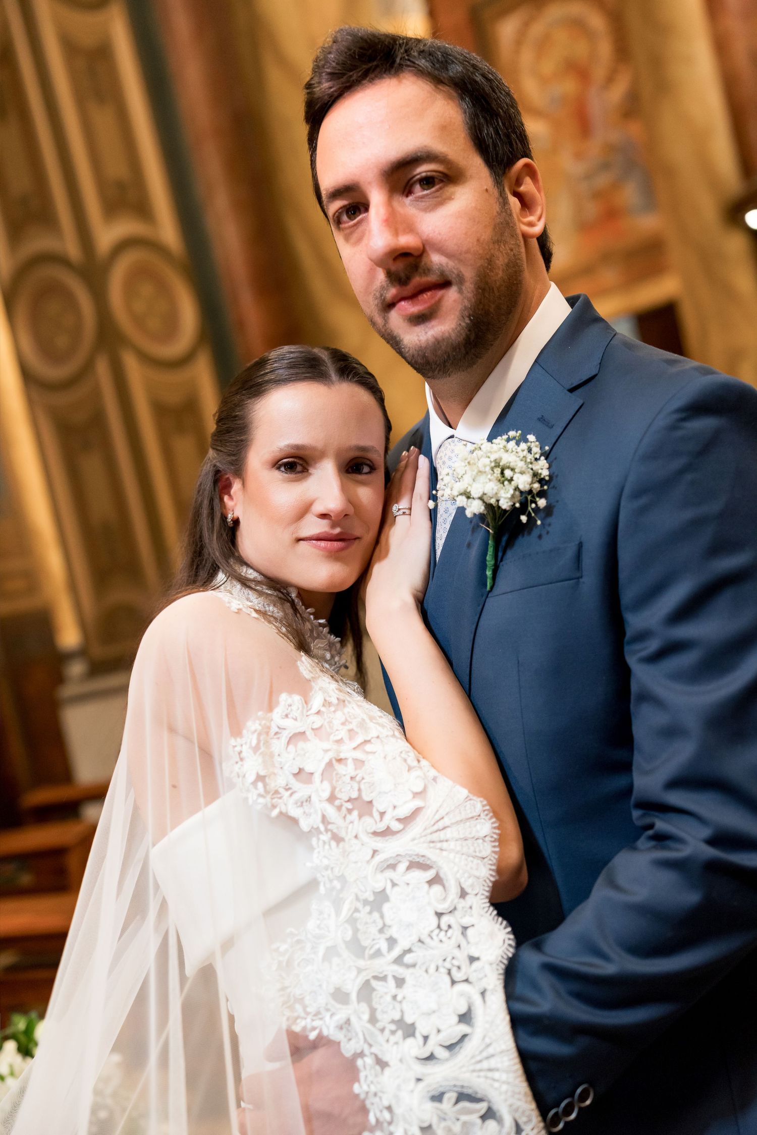 Bride and groom posing together in a church, bride in lace gown with veil, groom in navy suit with boutonniere.