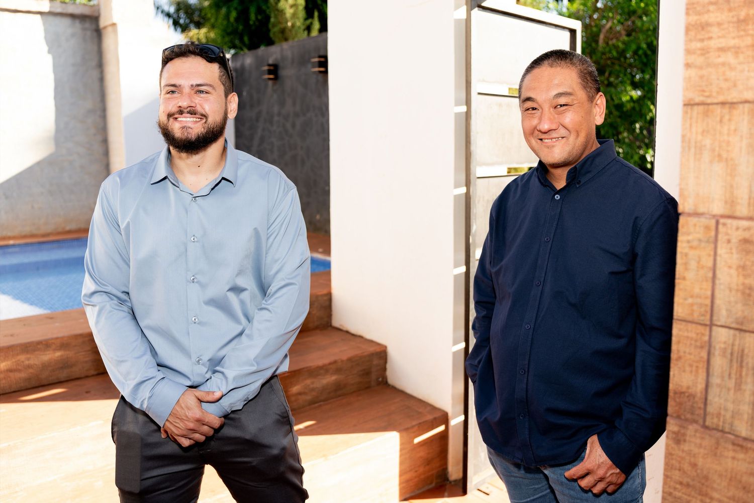 Two smiling men standing indoors near a pool area, wearing casual button-up shirts in a bright, modern home.