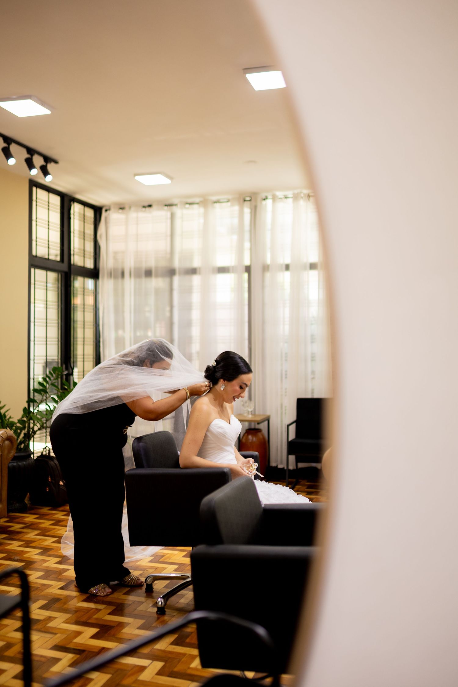 Bride seated in salon chair having veil adjusted by assistant in elegant room with herringbone floors.
