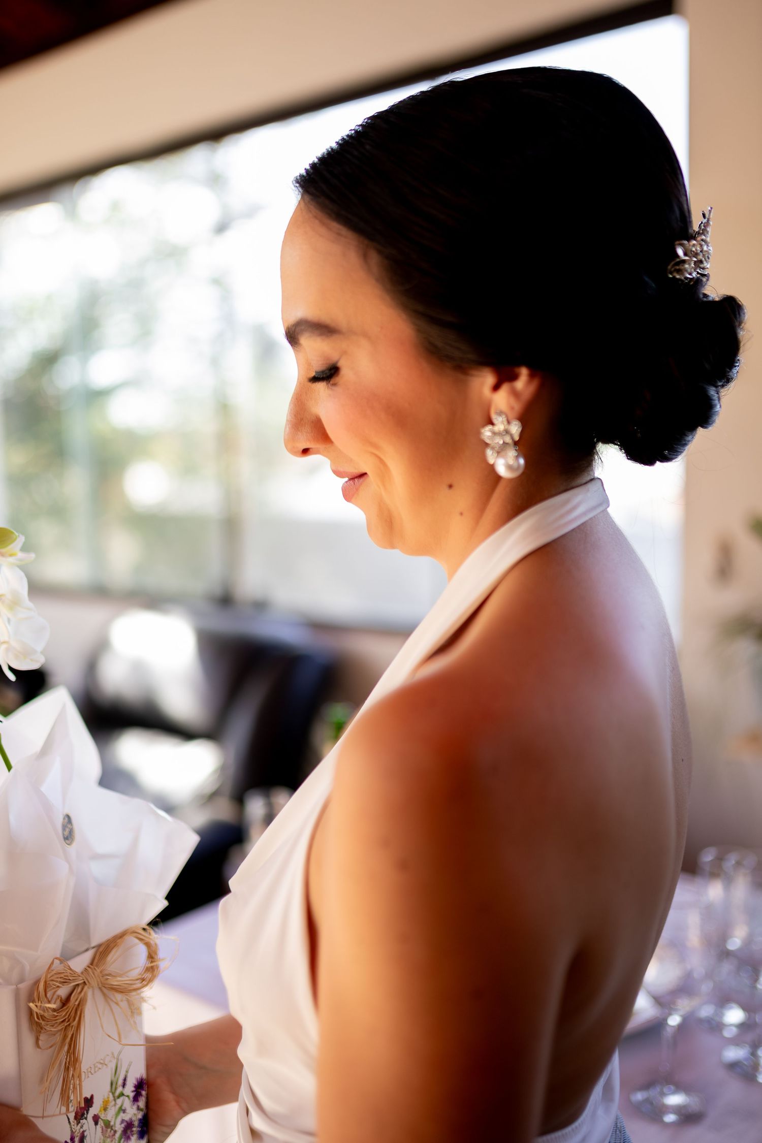 Smiling bride in white halter gown with pearl earrings and elegant updo holding a bouquet at her wedding reception.