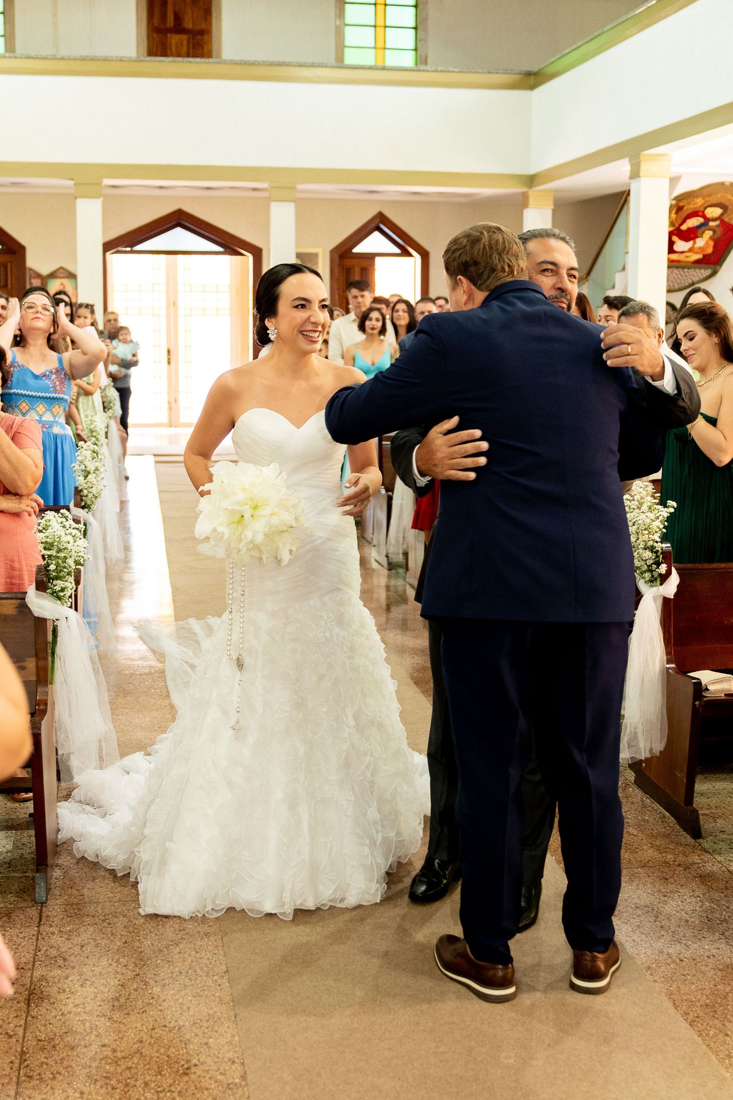 Bride in white gown smiling as man in navy suit embraces another guest during church wedding ceremony.