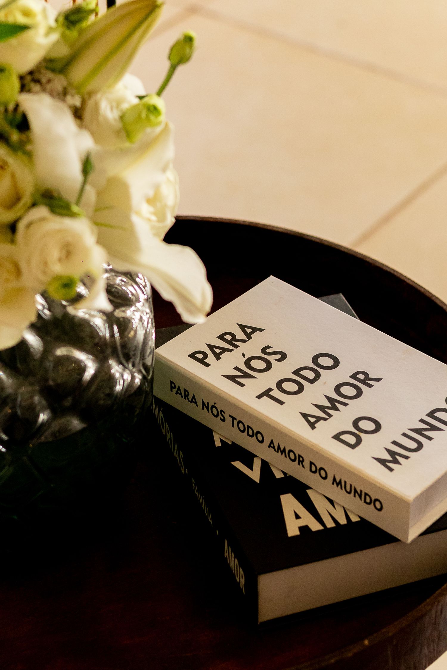 Books titled 'Para Nos Todo Amor Do Mundo' displayed on a dark tray beside white flowers.