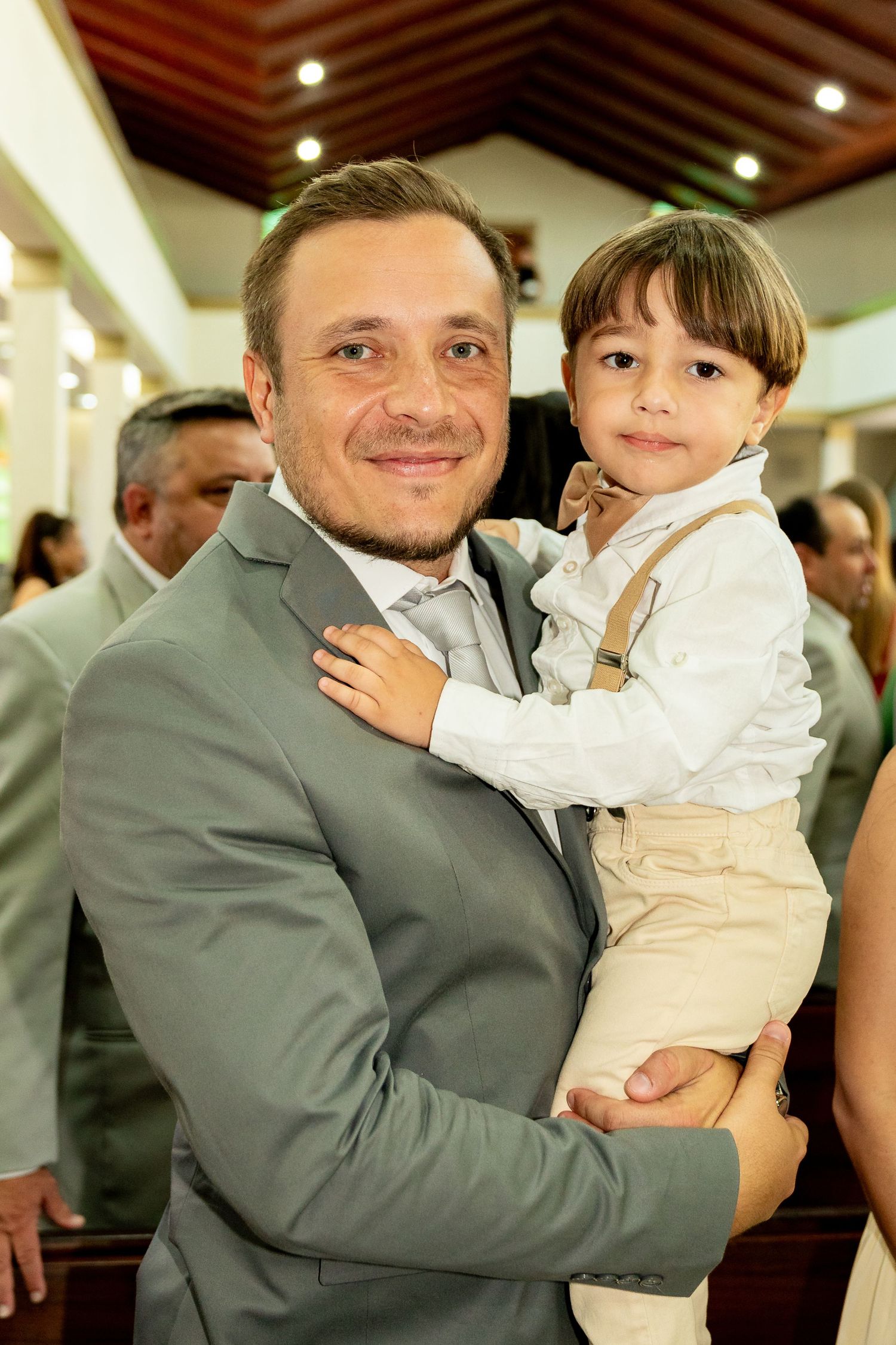 Man in gray suit holding young boy in white shirt and suspenders at a formal event.