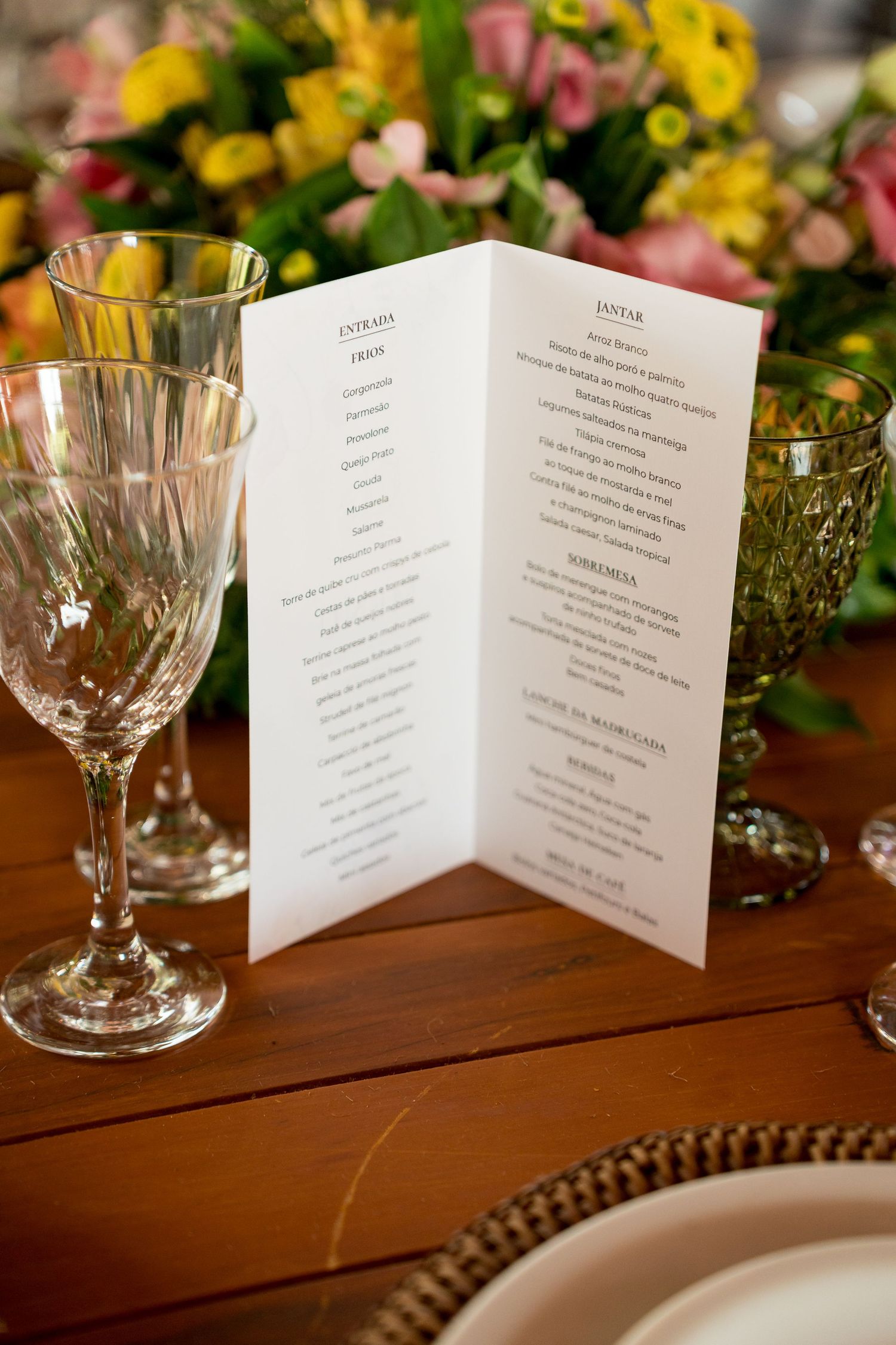 Elegant folded wedding menu card on wooden table with crystal wine glasses and colorful floral centerpiece.