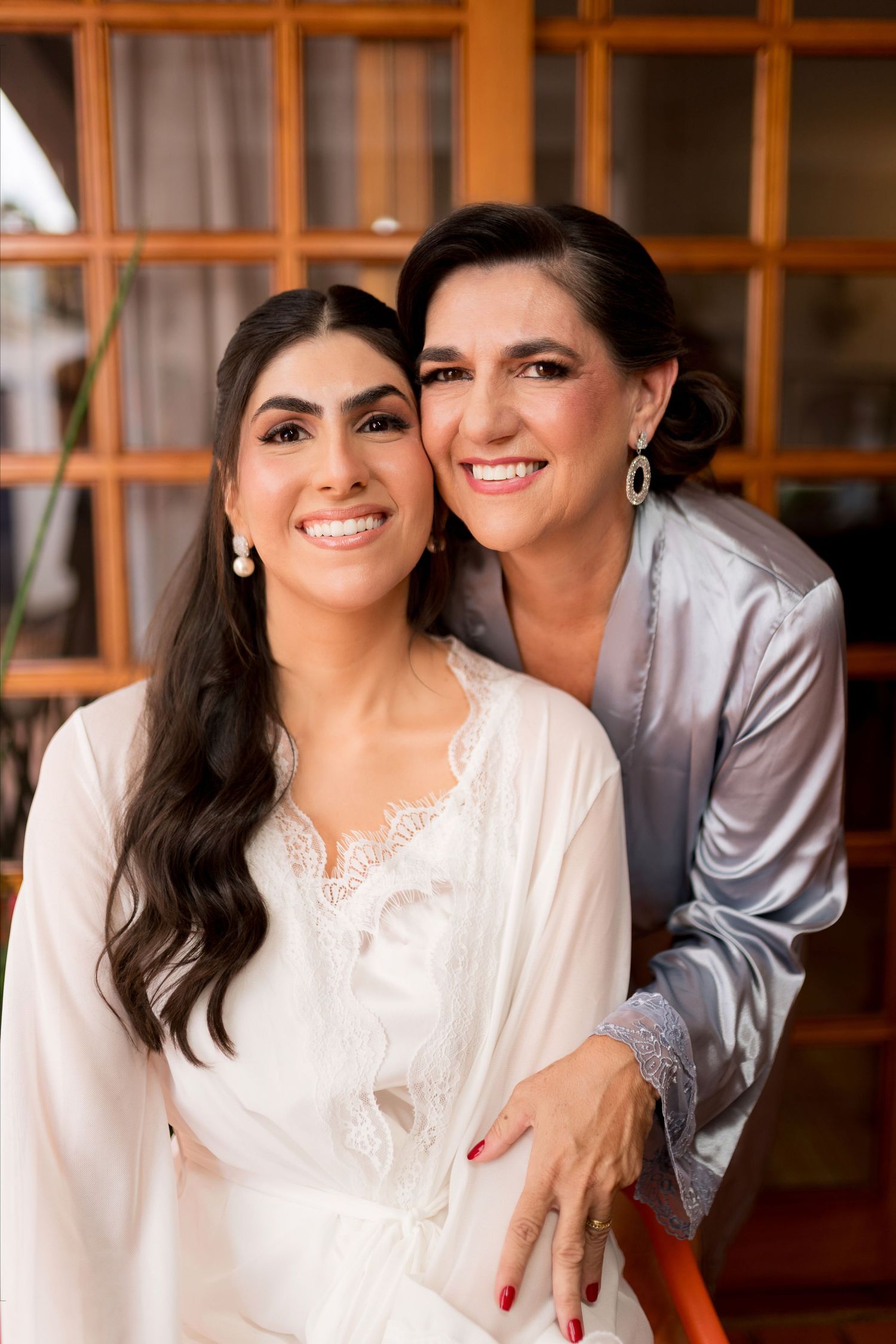 Two women in satin robes smile together, one in white lace bridal robe, posing indoors before a wedding ceremony.