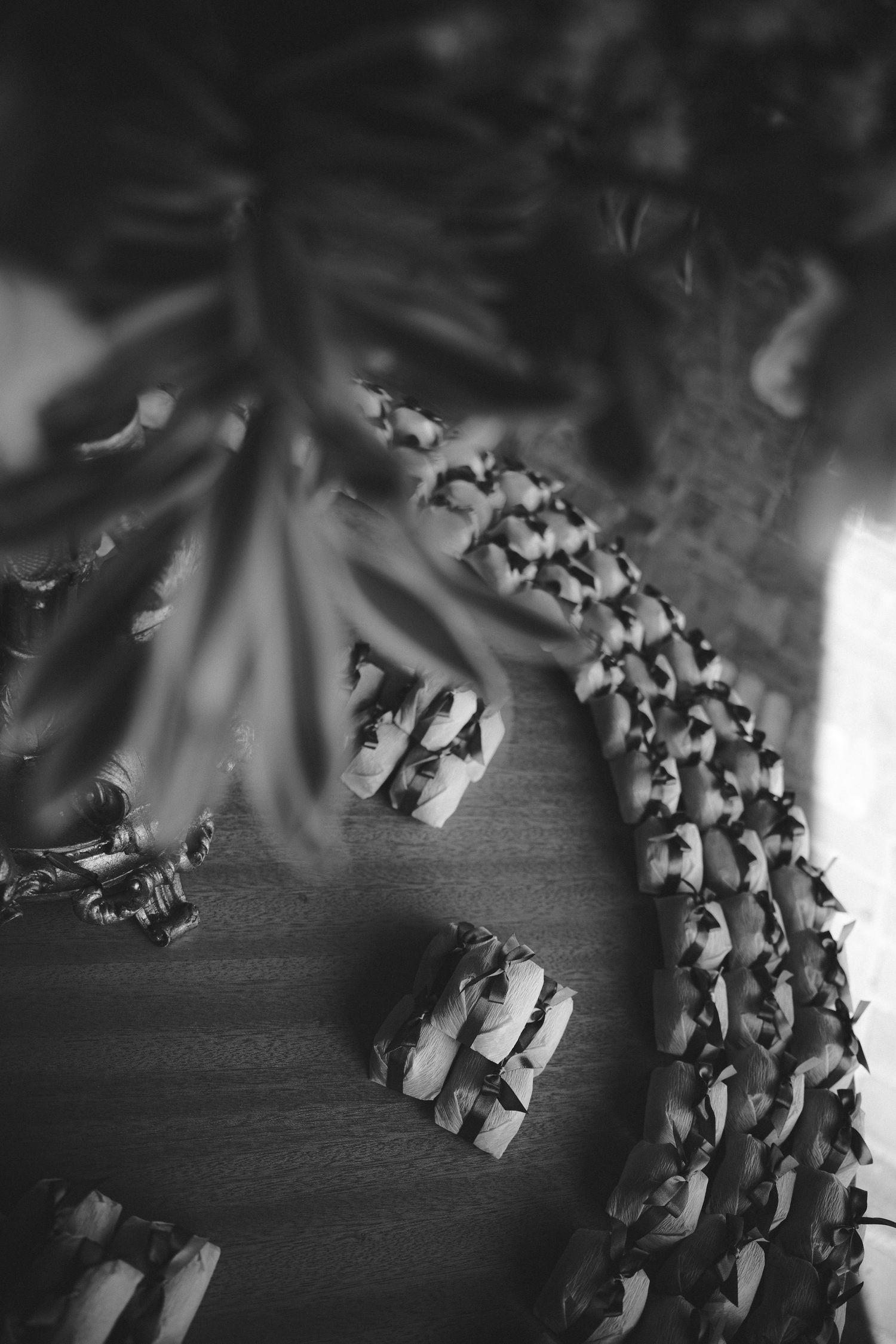 Black and white photo of floral garlands and small gift bundles arranged on a wooden surface with tropical leaves overhead.