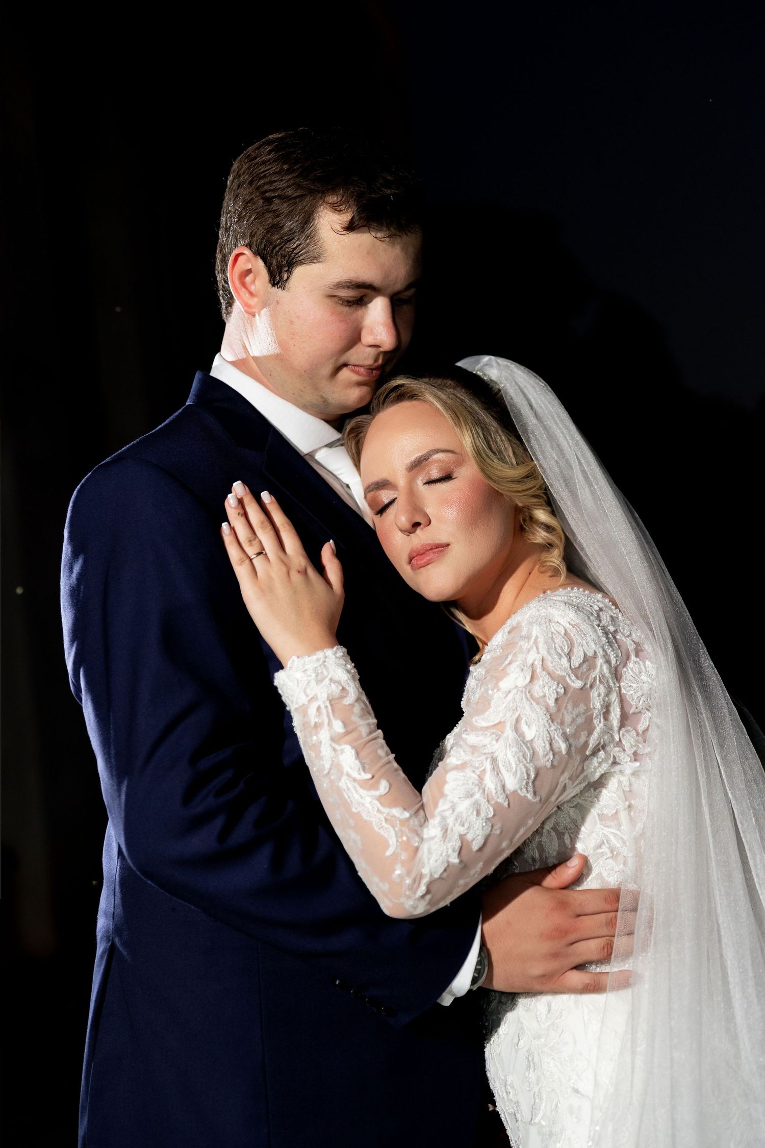 Bride and groom share a tender embrace, she in lace wedding gown with veil, he in navy suit, against dark background.
