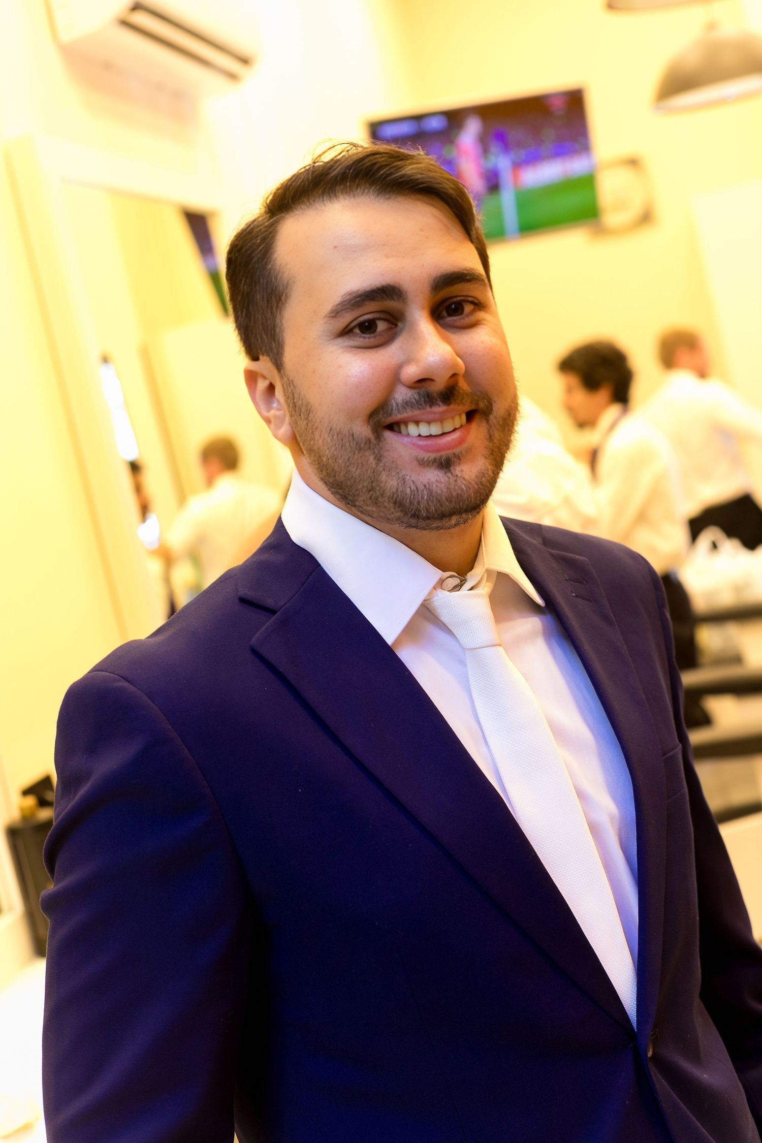 Smiling man in navy blue suit and white tie poses in a barbershop with TV in background.