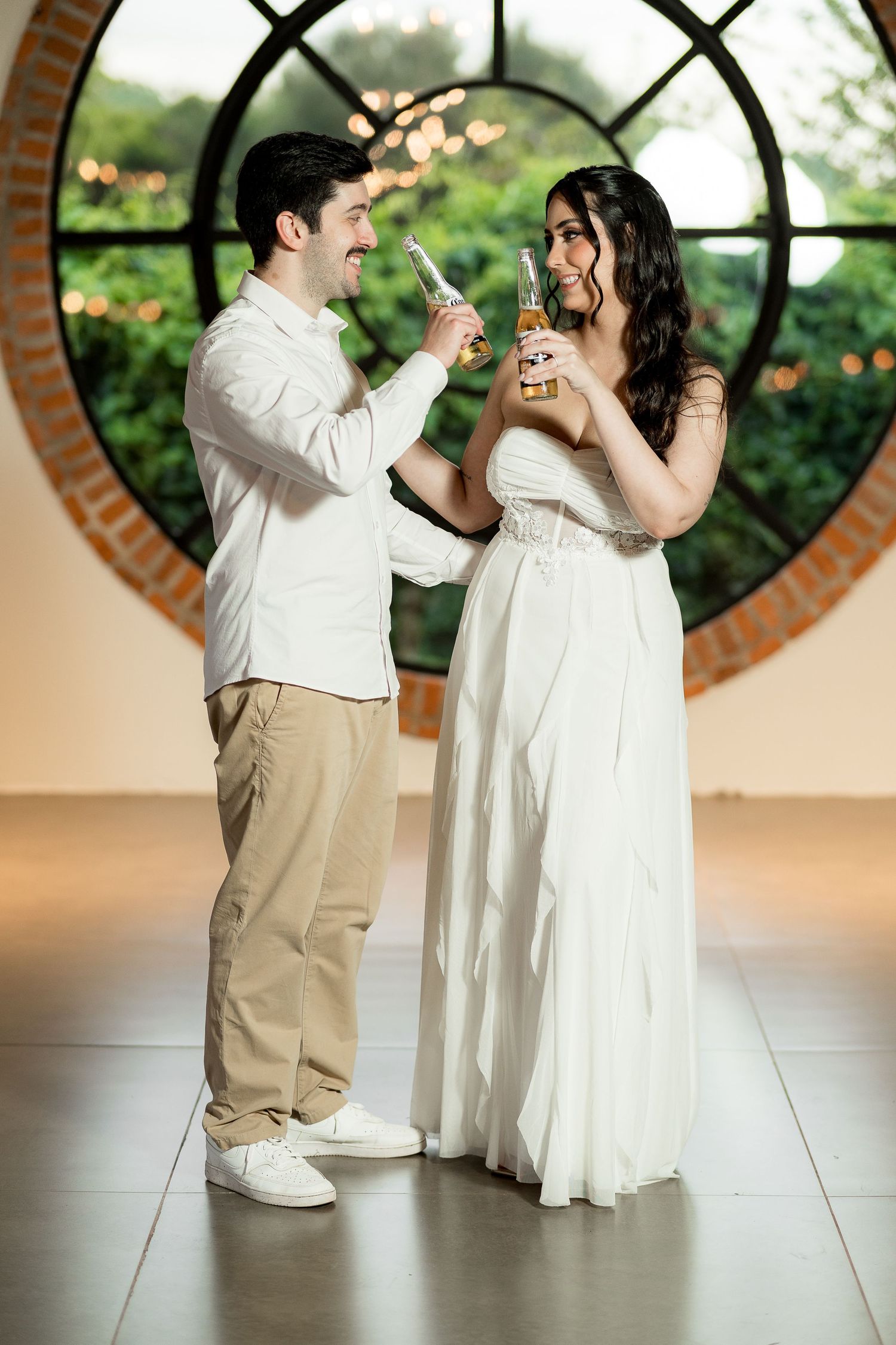 A couple in wedding attire toasting with beer bottles in front of a round window.