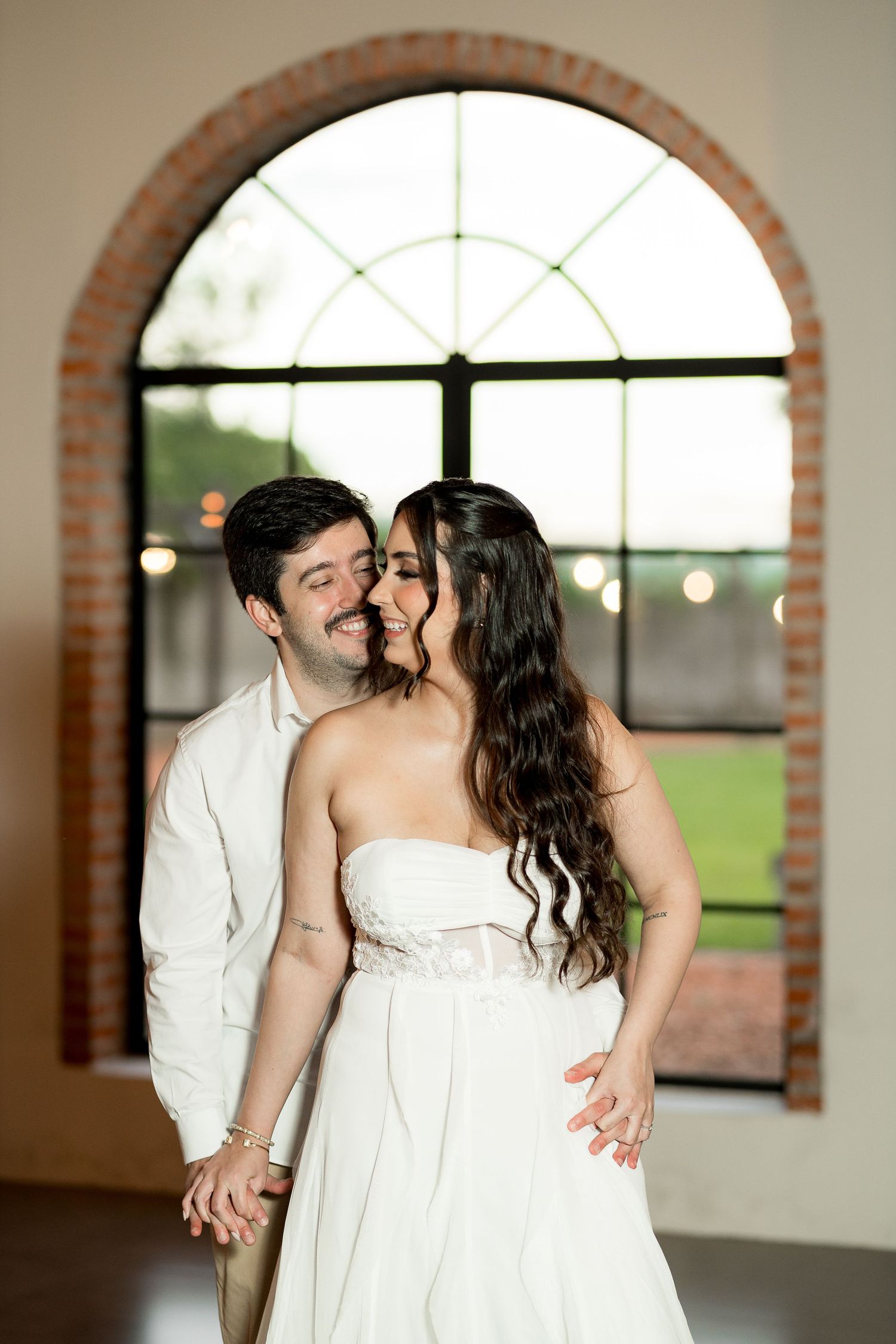 Couple in white wedding attire sharing a romantic kiss in front of an arched brick window.
