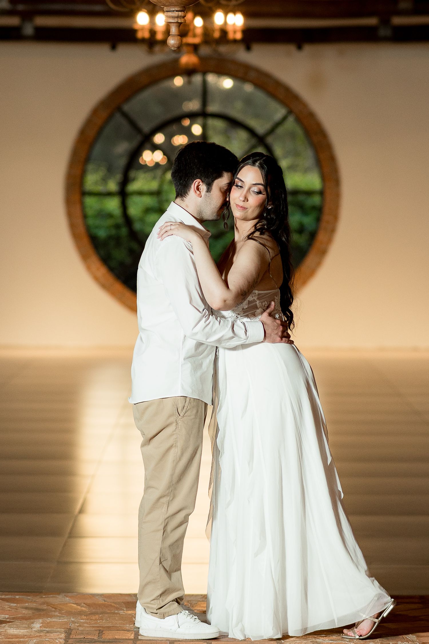 Couple sharing a romantic embrace in elegant white attire before a grand circular window in a warmly lit venue.