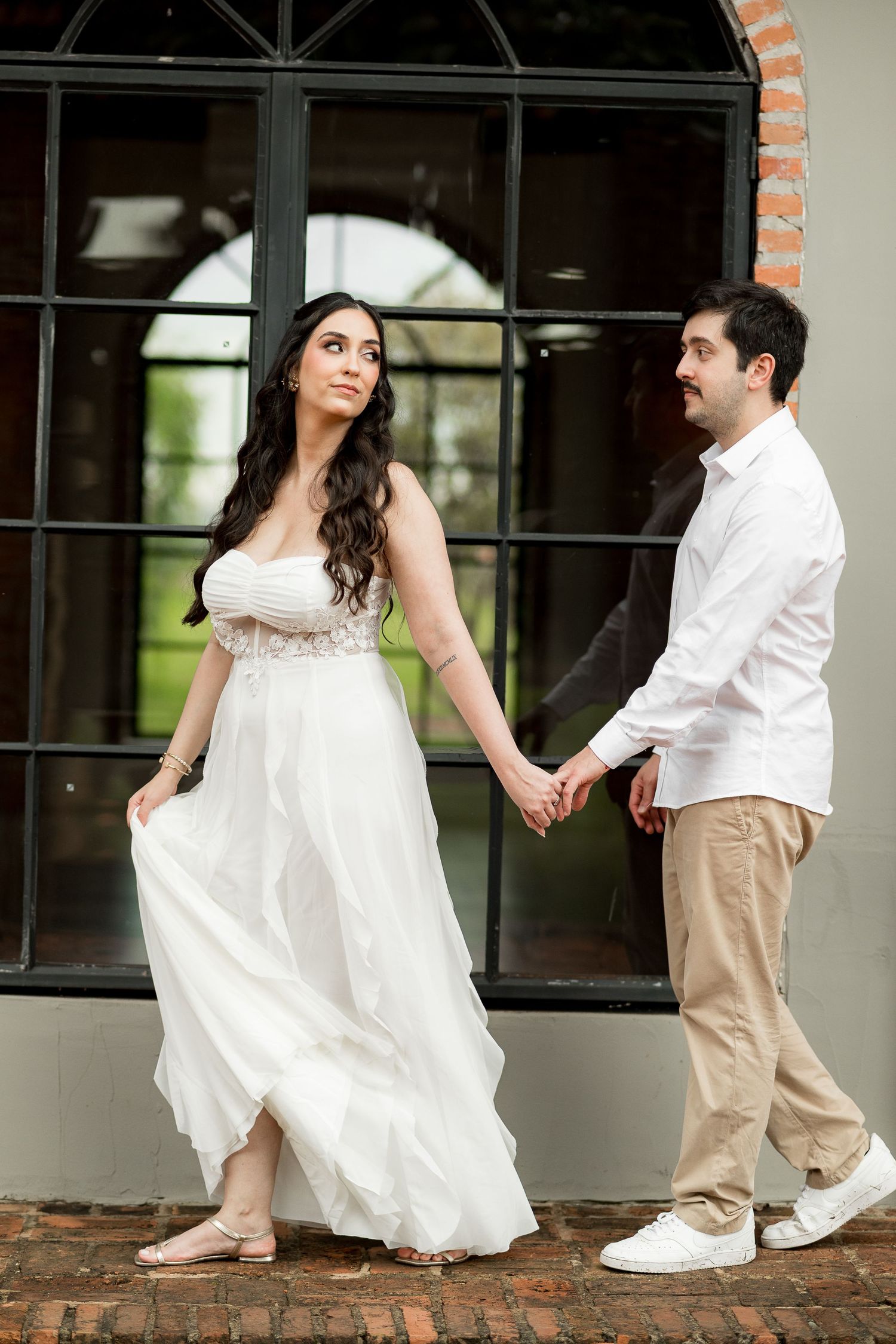 A couple in wedding attire holds hands outdoors, the bride in a flowing white gown and groom in white shirt and khakis.