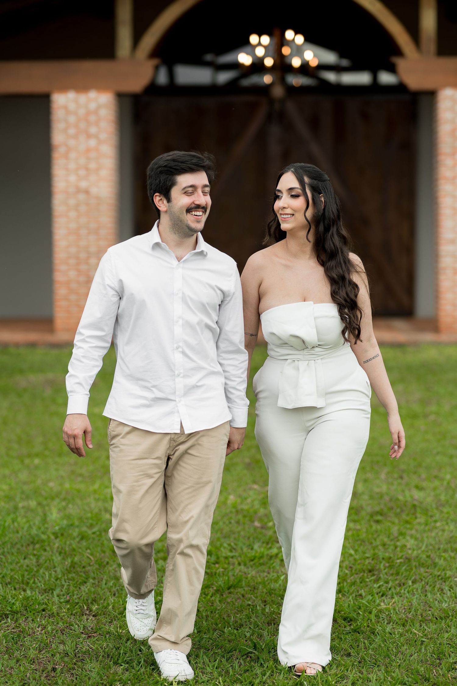 A smiling couple walks on green grass, man in white shirt and khakis, woman in white strapless jumpsuit, barn background.