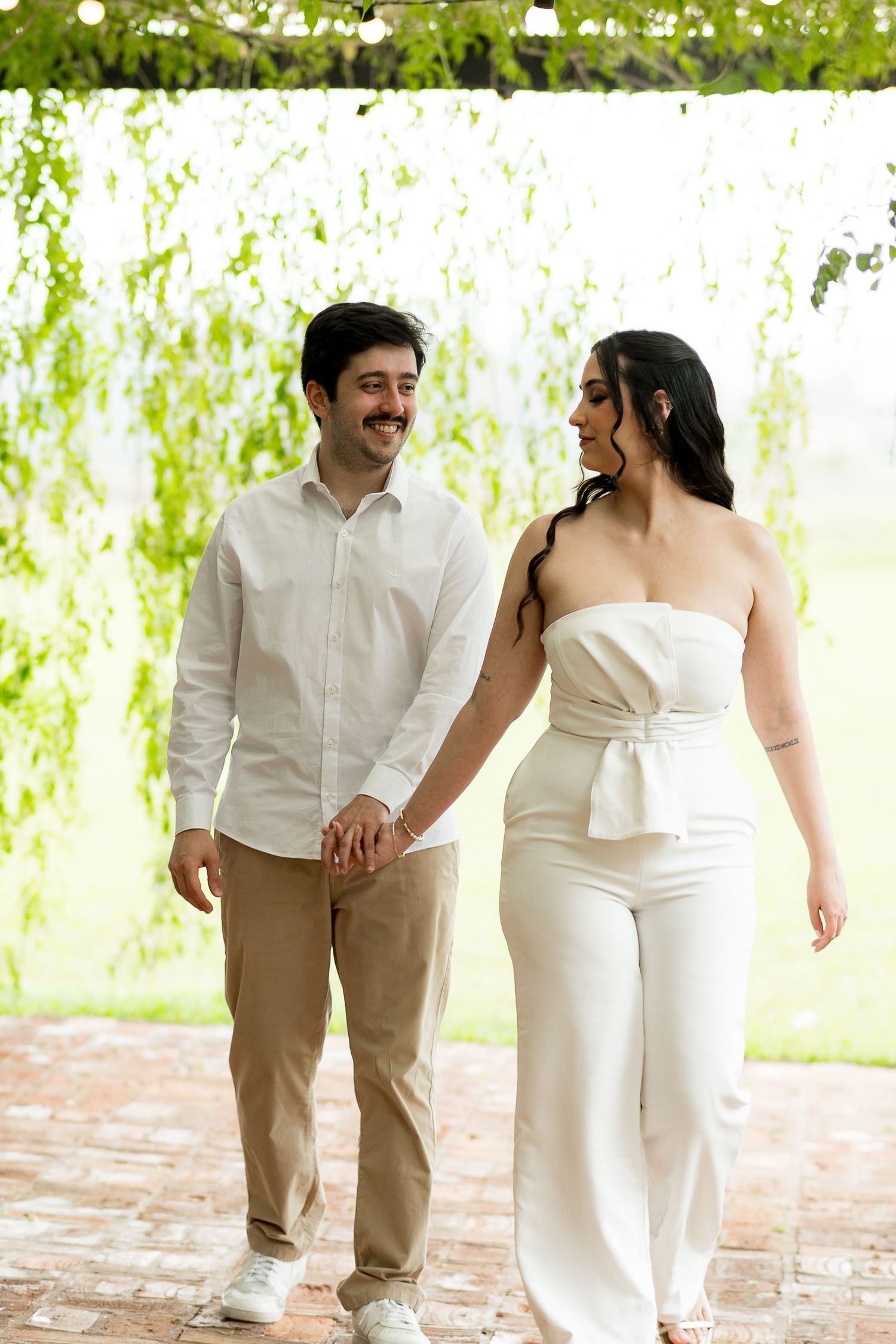 A smiling couple in white and beige outfits hold hands while walking under lush green weeping willow branches.