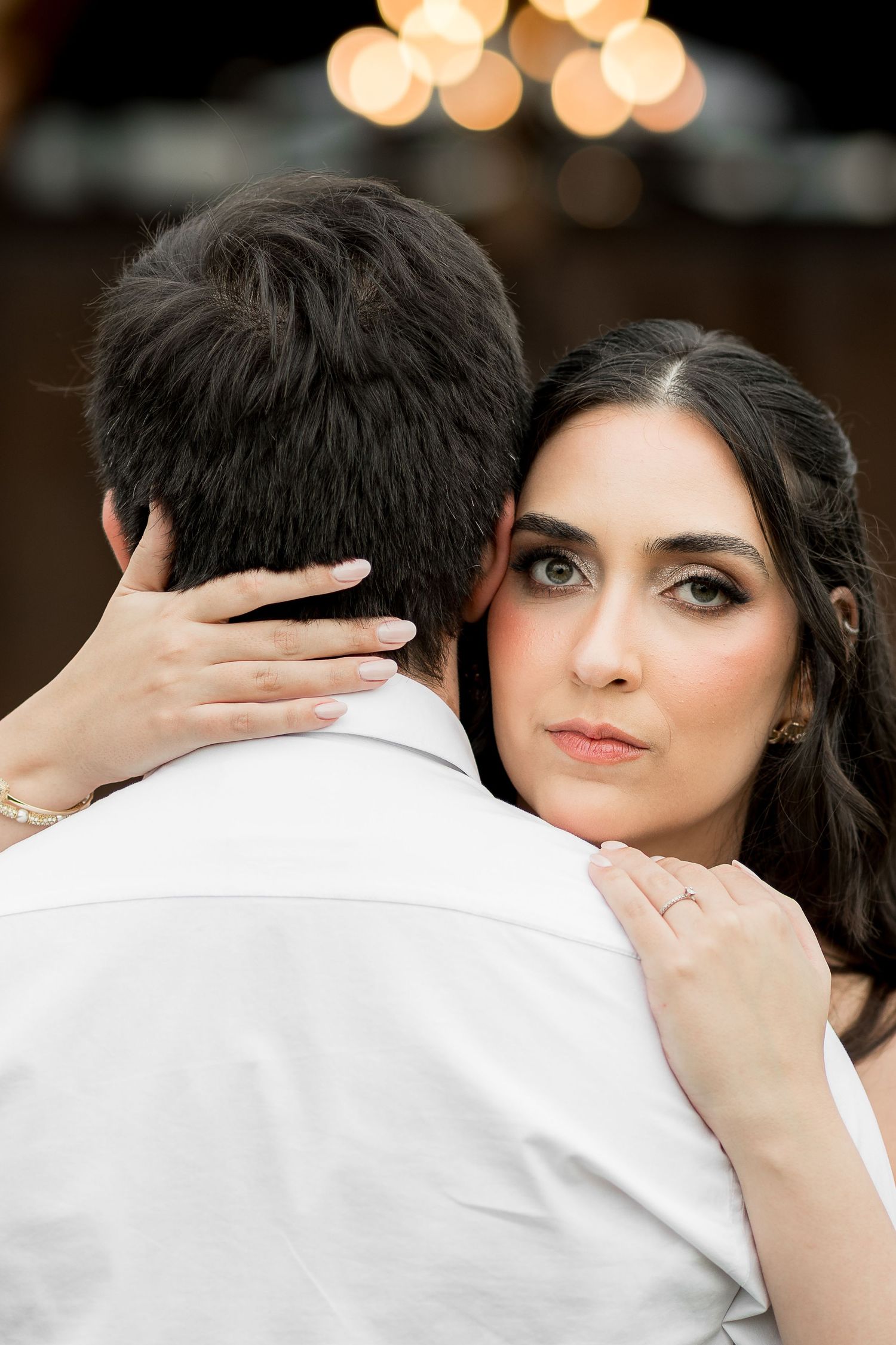 Bride with dark hair and smoky eye makeup embracing groom in white shirt at romantic event.