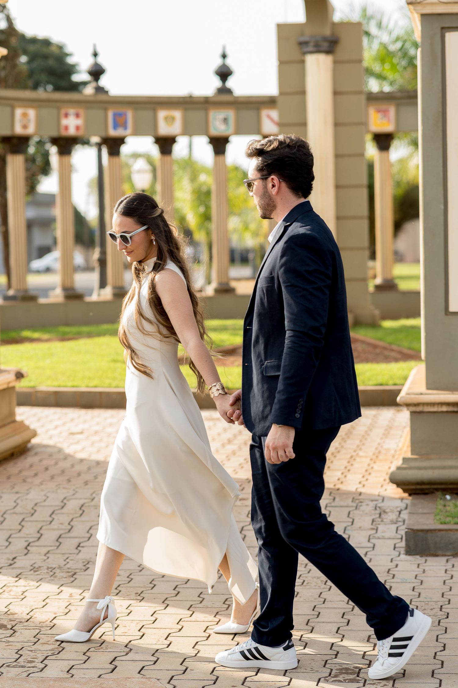 Elegant couple holding hands walking through a columned outdoor venue, woman in white dress and man in navy suit.