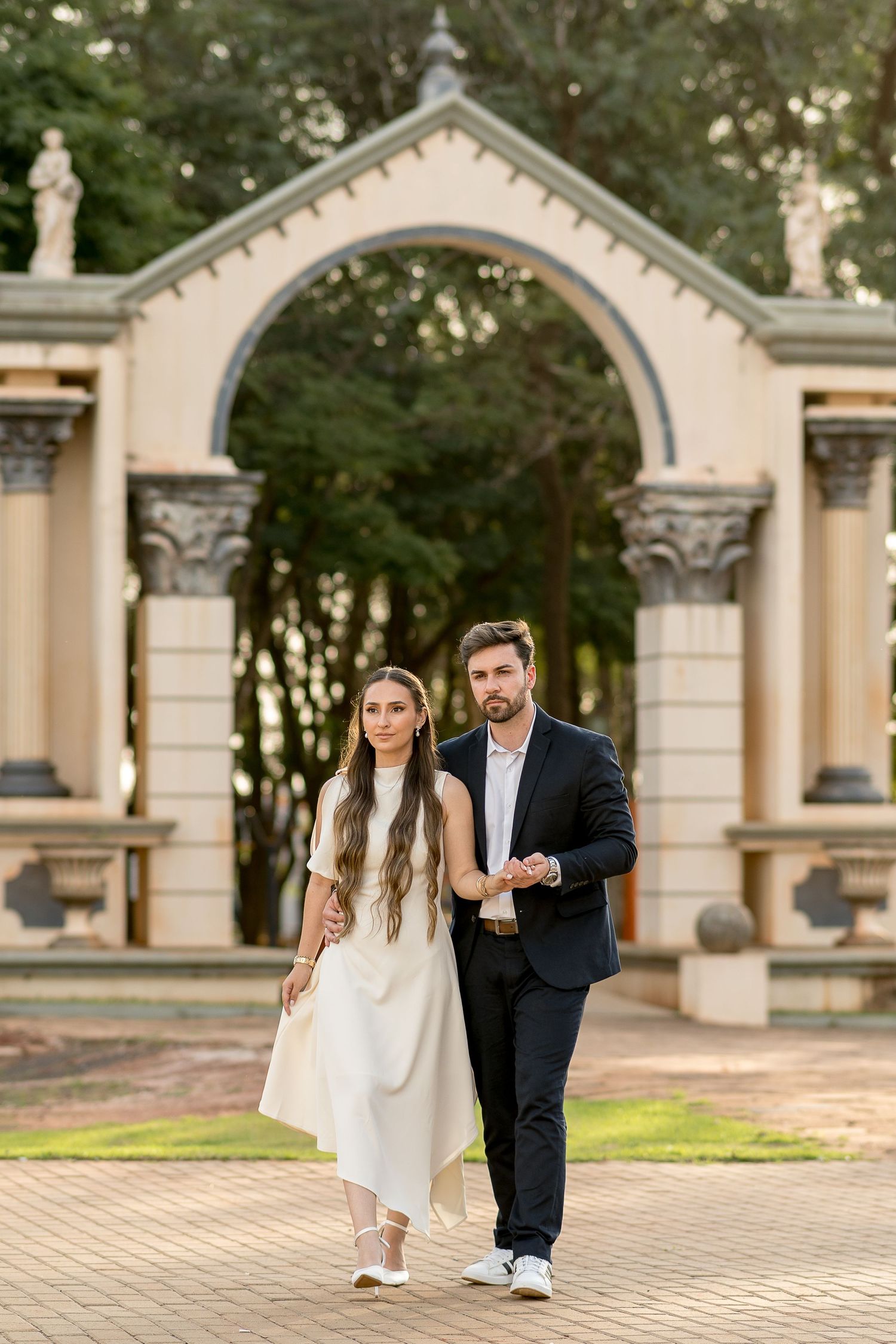 Elegant couple walking through ornate stone archway in lush garden, woman in white dress, man in dark suit.