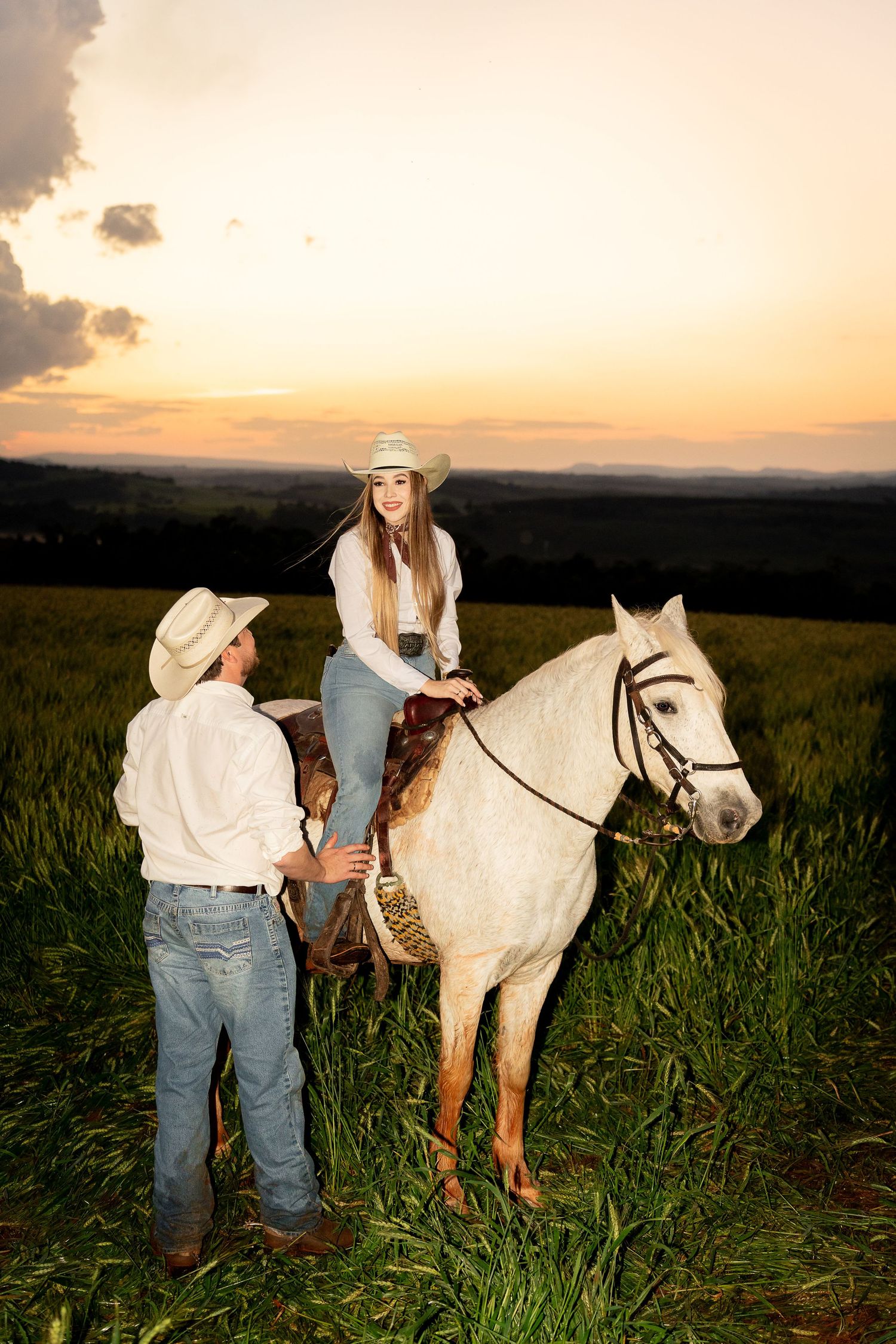 A woman in a cowboy hat rides a white horse in a green field at sunset, while a man in a white shirt stands beside her.