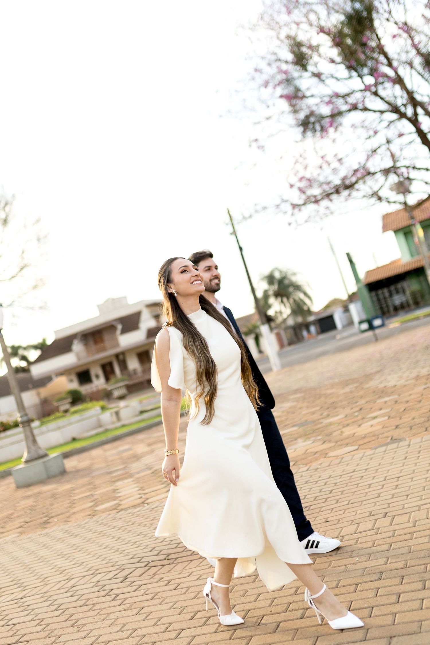 A smiling bride in a white dress and groom in a dark suit walk together on a brick plaza outdoors.