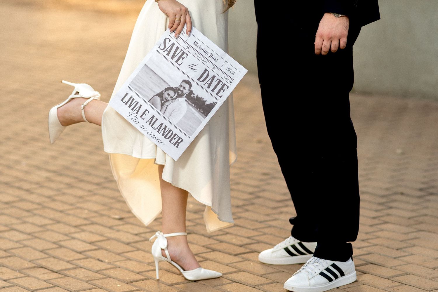 Couple holding a Save the Date newspaper announcement, bride in white heels and groom in Adidas sneakers on brick pavement.