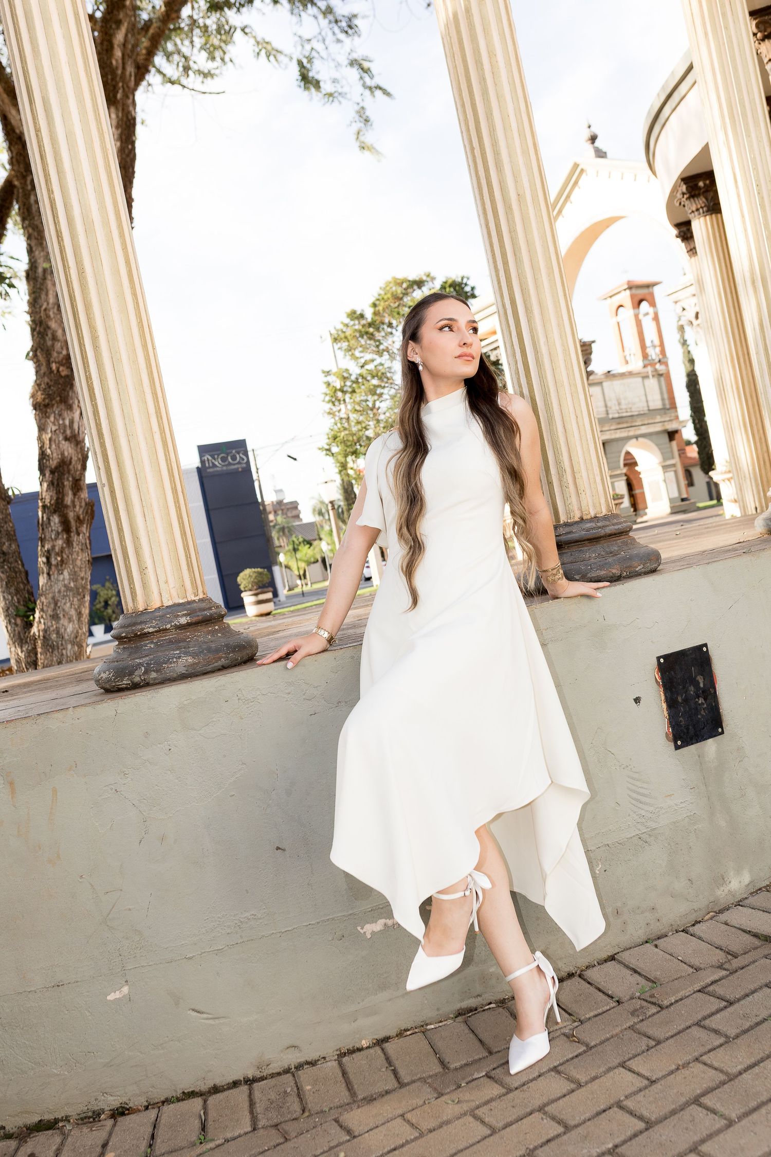 Young woman in white asymmetric dress and heels posing by classical columns in an outdoor urban setting.