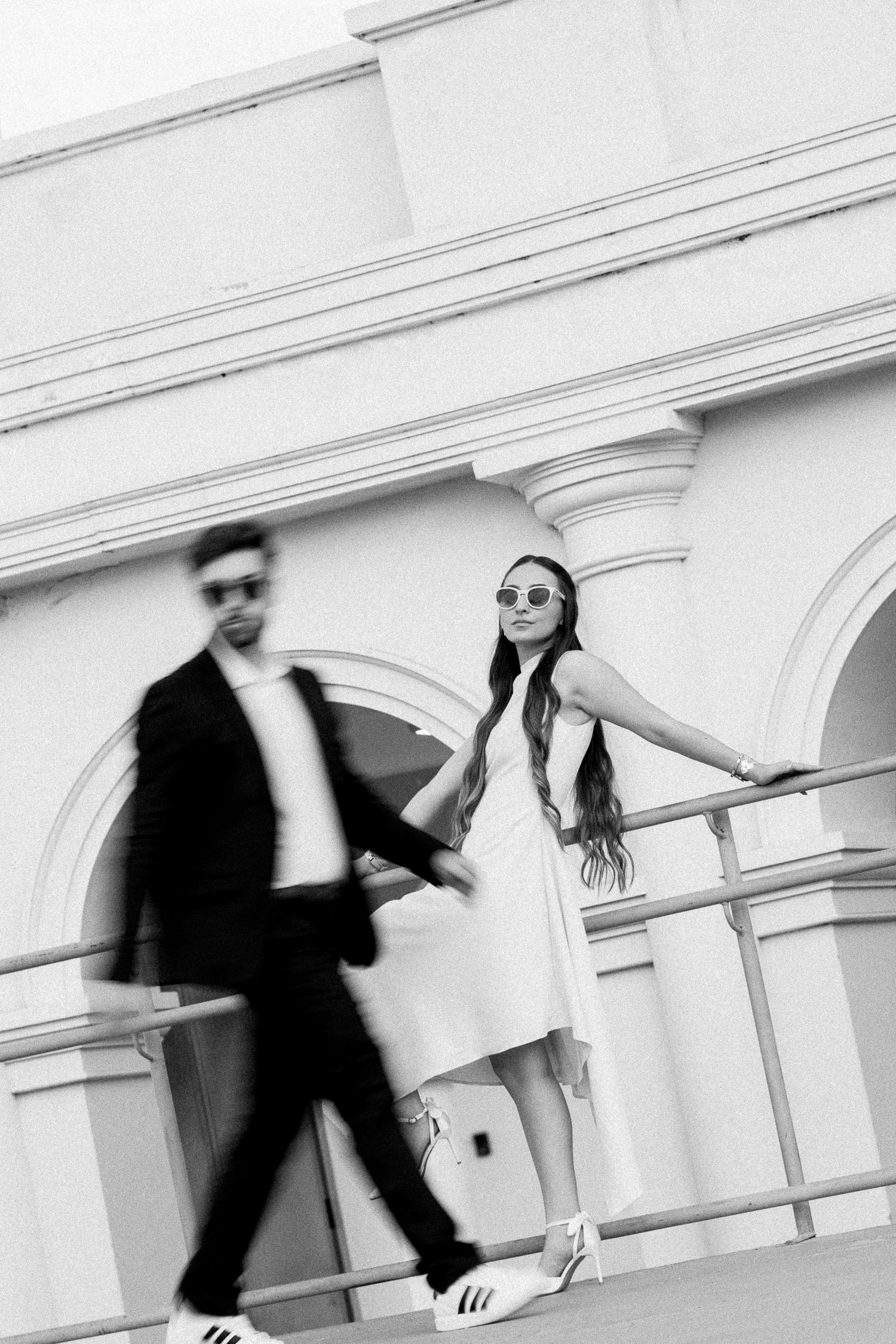 Black and white photo of a couple holding hands near ornate building columns, woman in white dress and sunglasses.