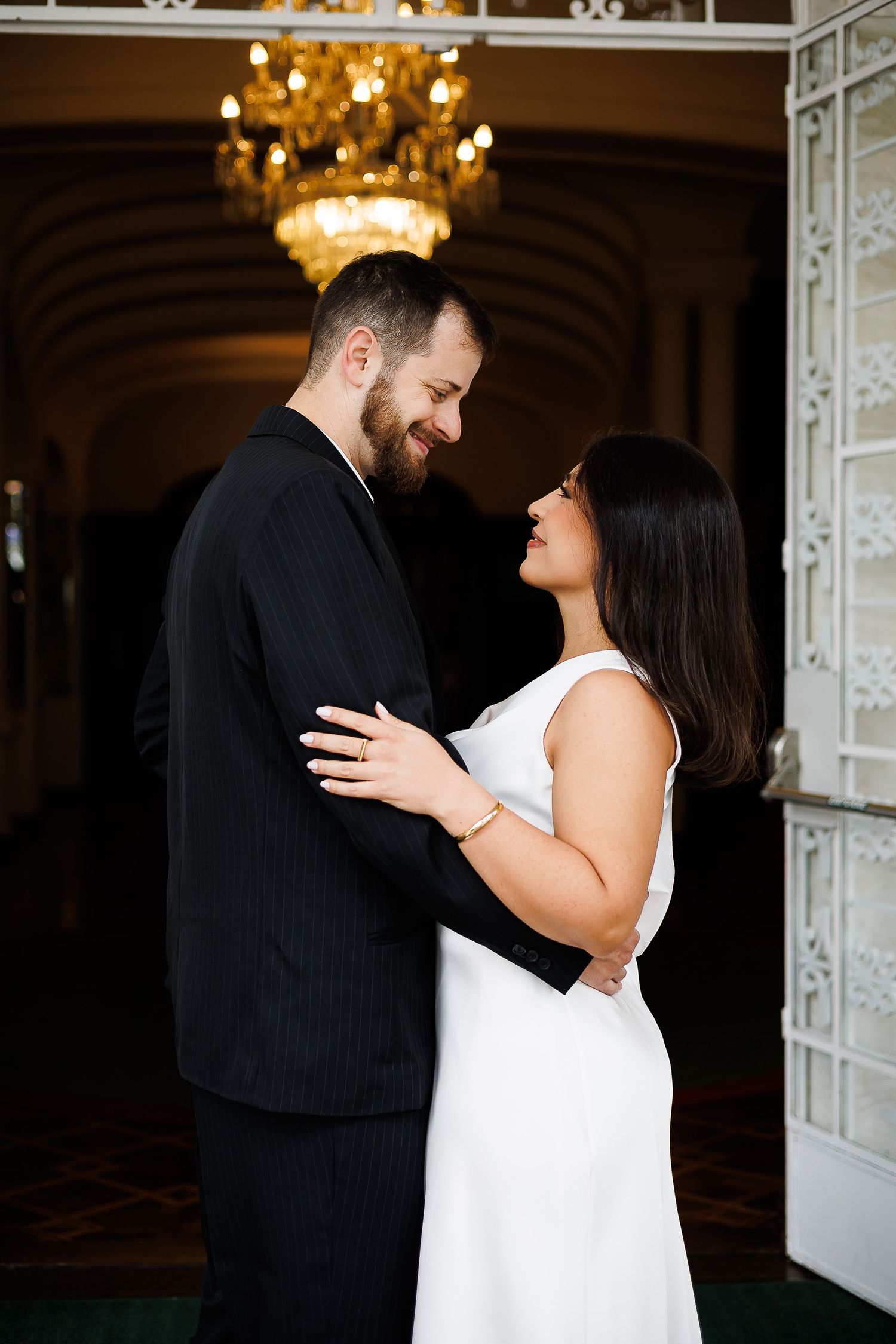 Couple in wedding attire embracing under a chandelier in an elegant venue.