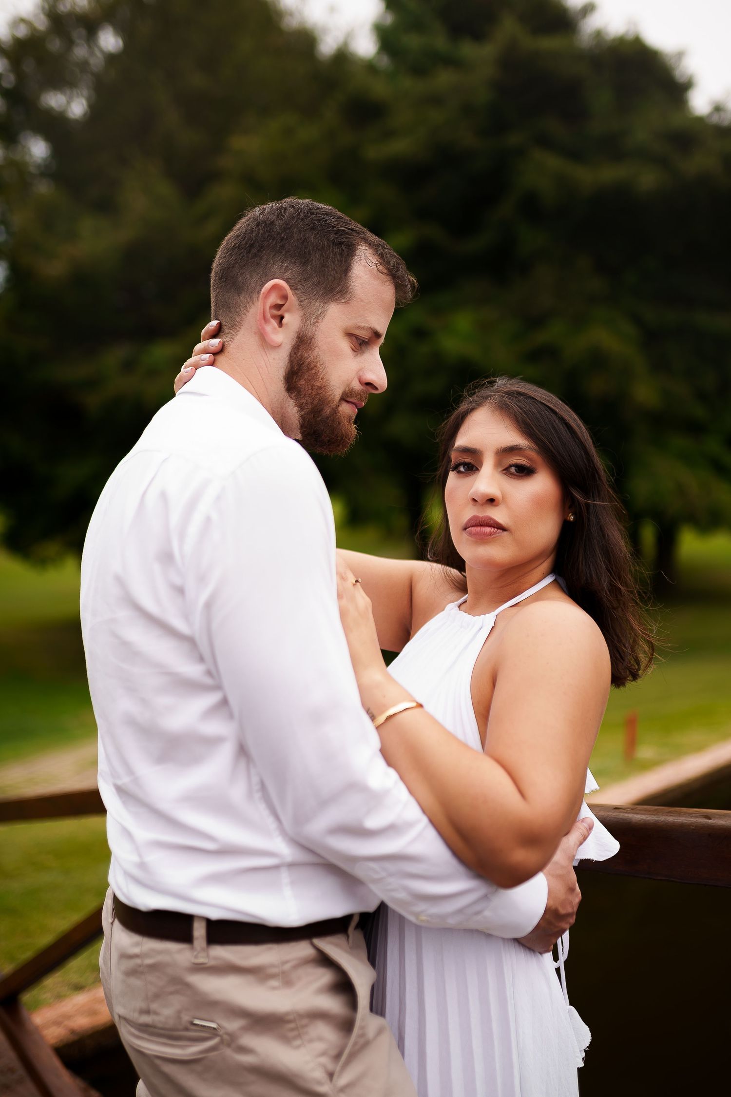 Couple embracing outdoors, woman in white dress looks at camera while man in white shirt holds her in a lush green park.