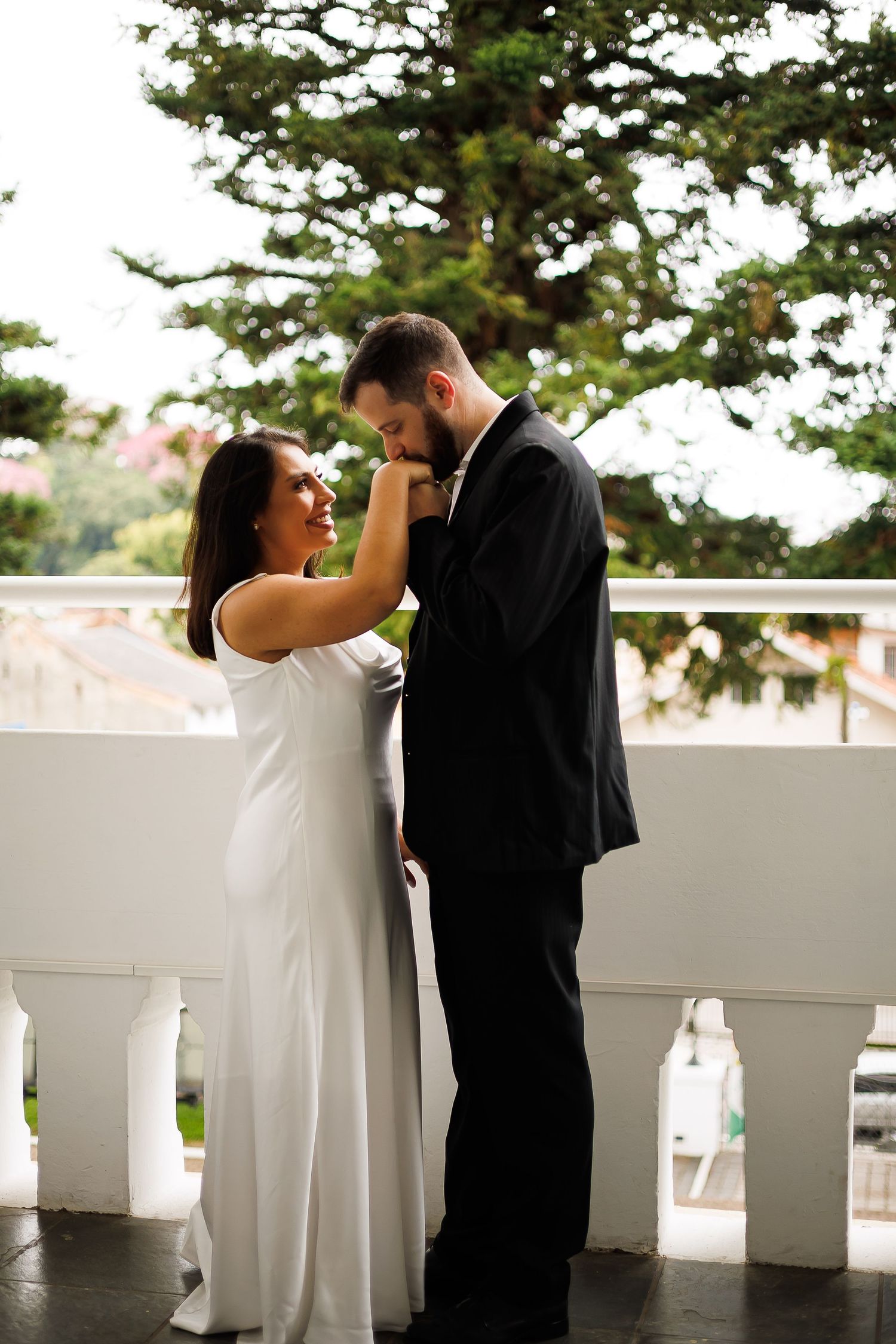 A bride in white gown and groom in black suit share a romantic moment on a white balcony outdoors.