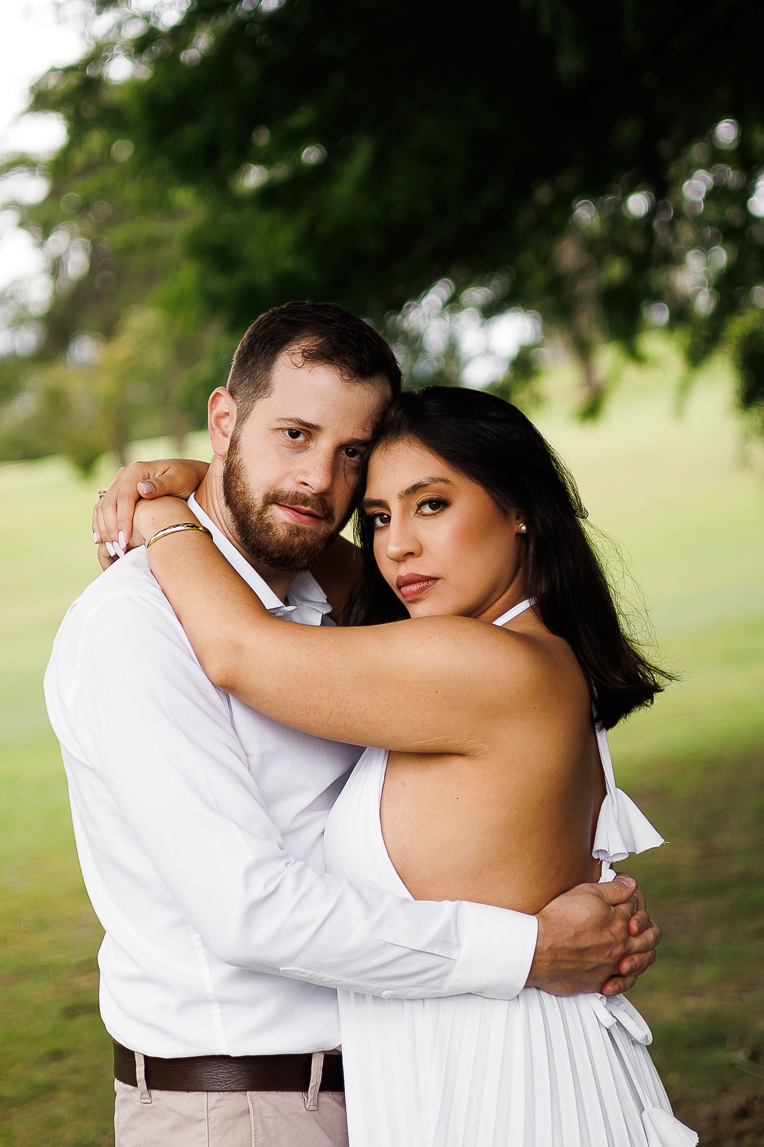 Couple in white outfits embracing outdoors, man with beard and woman with dark hair, green park background.