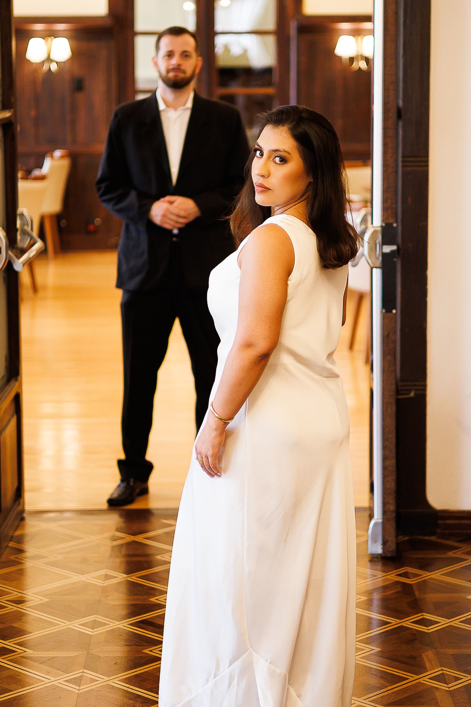 Bride in white gown looks back at groom in black suit standing in elegant room with ornate flooring.