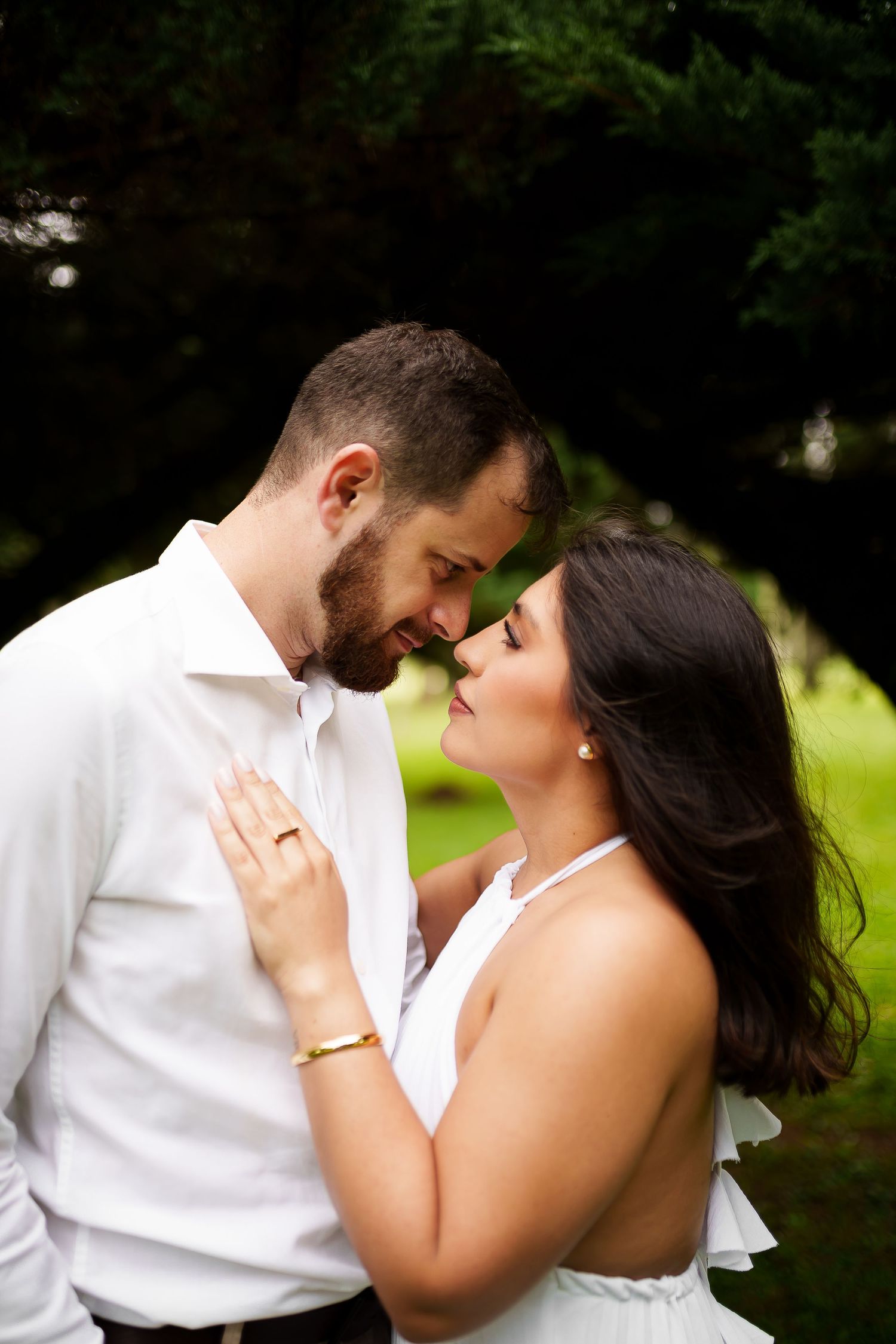 Romantic couple in white outfits sharing an intimate moment outdoors, touching foreheads in a lush green garden setting.