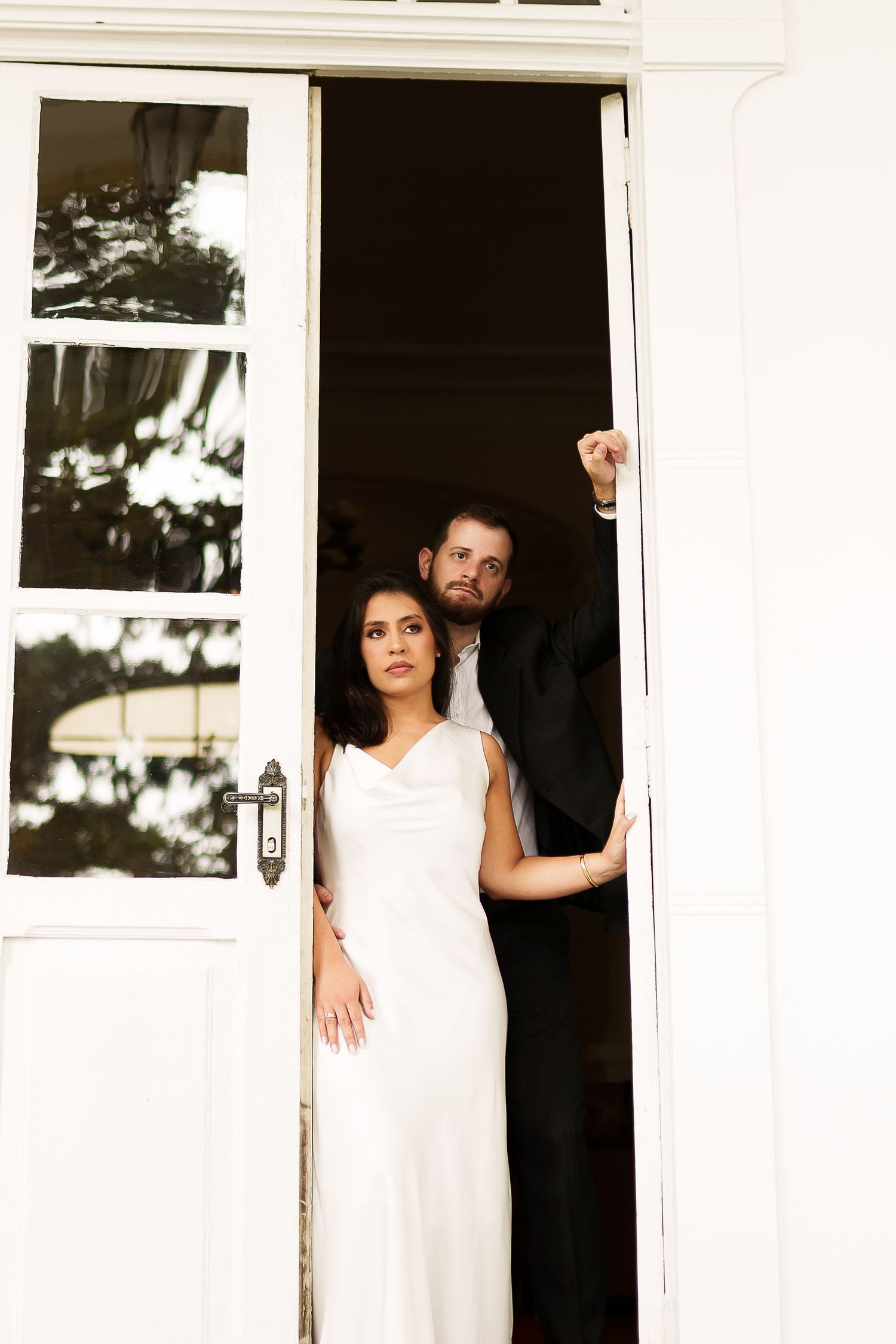 Bride in white gown and groom in black suit stand in elegant white double doorway for wedding portrait.