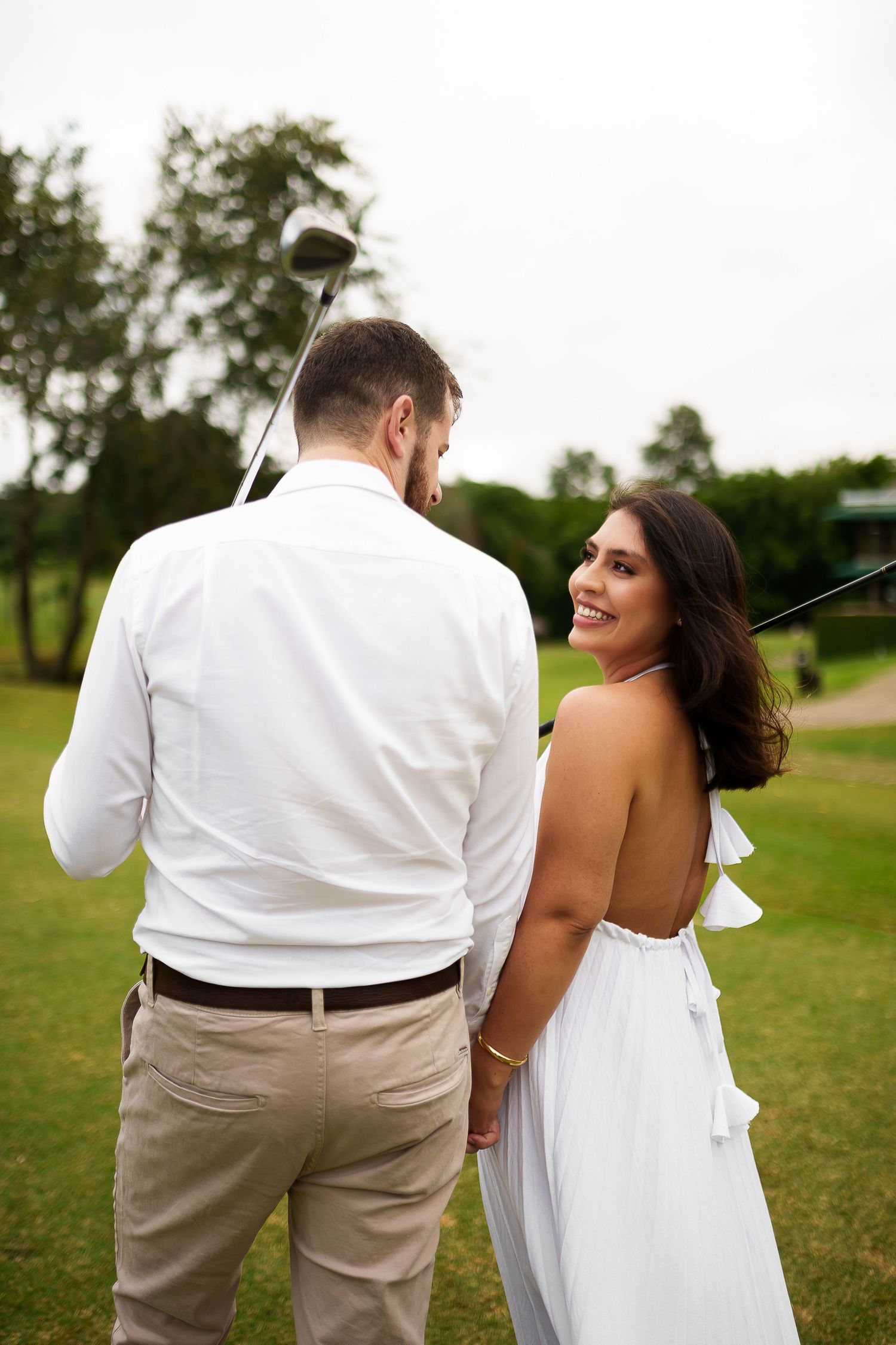 Couple holding hands on a golf course, woman in white backless dress smiling, man in white shirt holding a golf club.