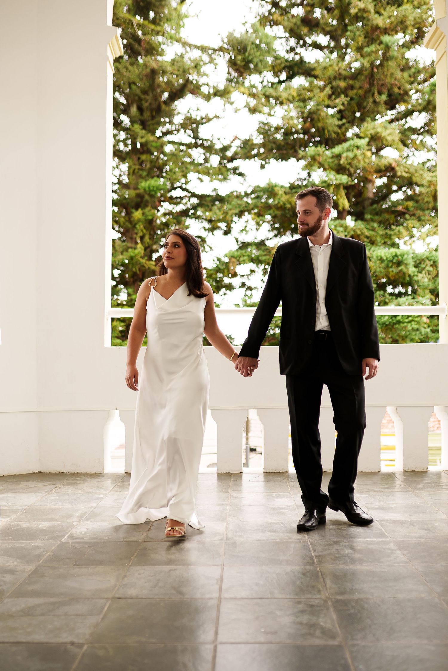 Bride in white gown and groom in black suit hold hands while walking on elegant white columned porch.