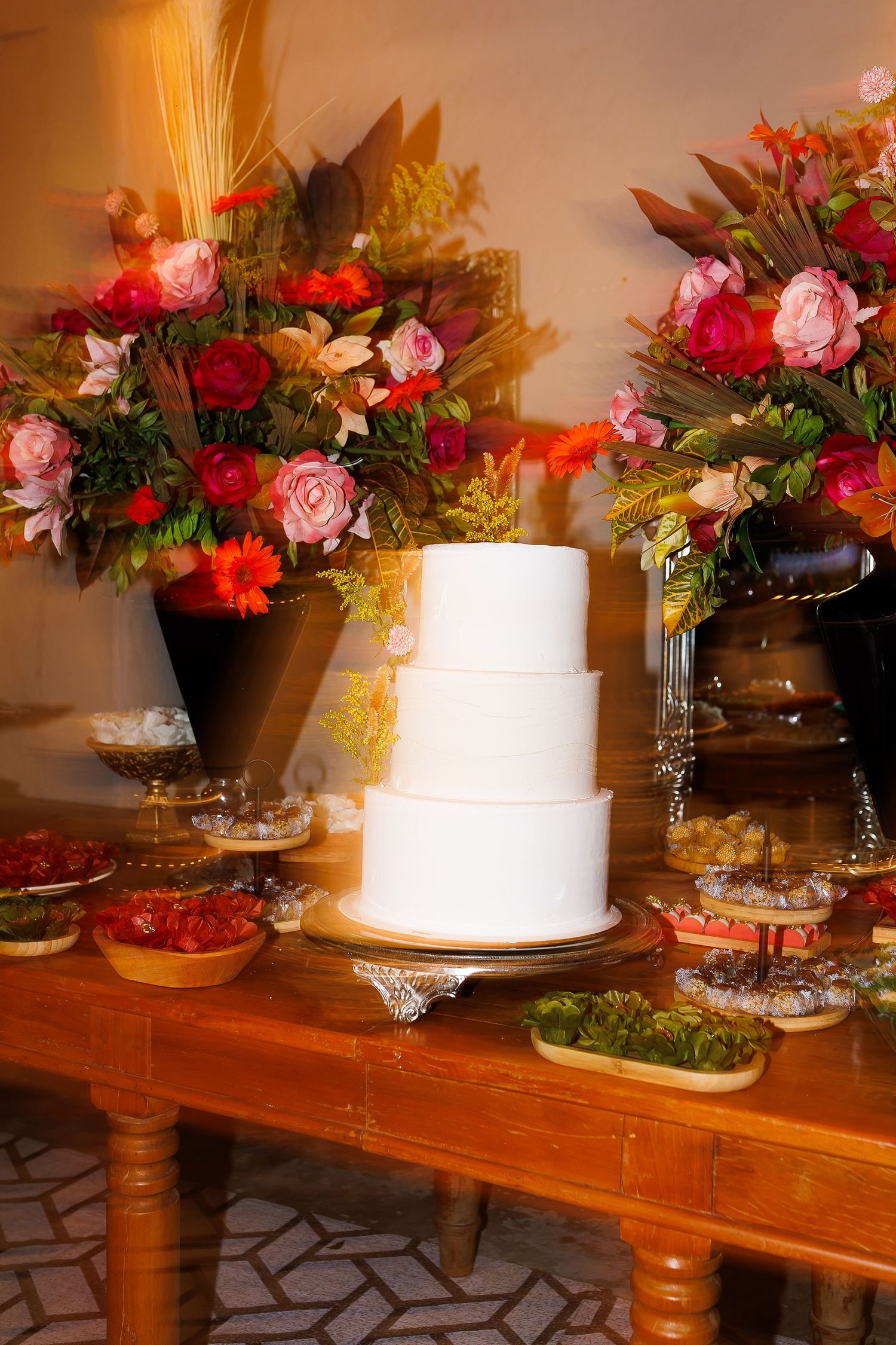 Elegant white three-tier wedding cake on wooden table with colorful floral arrangements and assorted desserts.