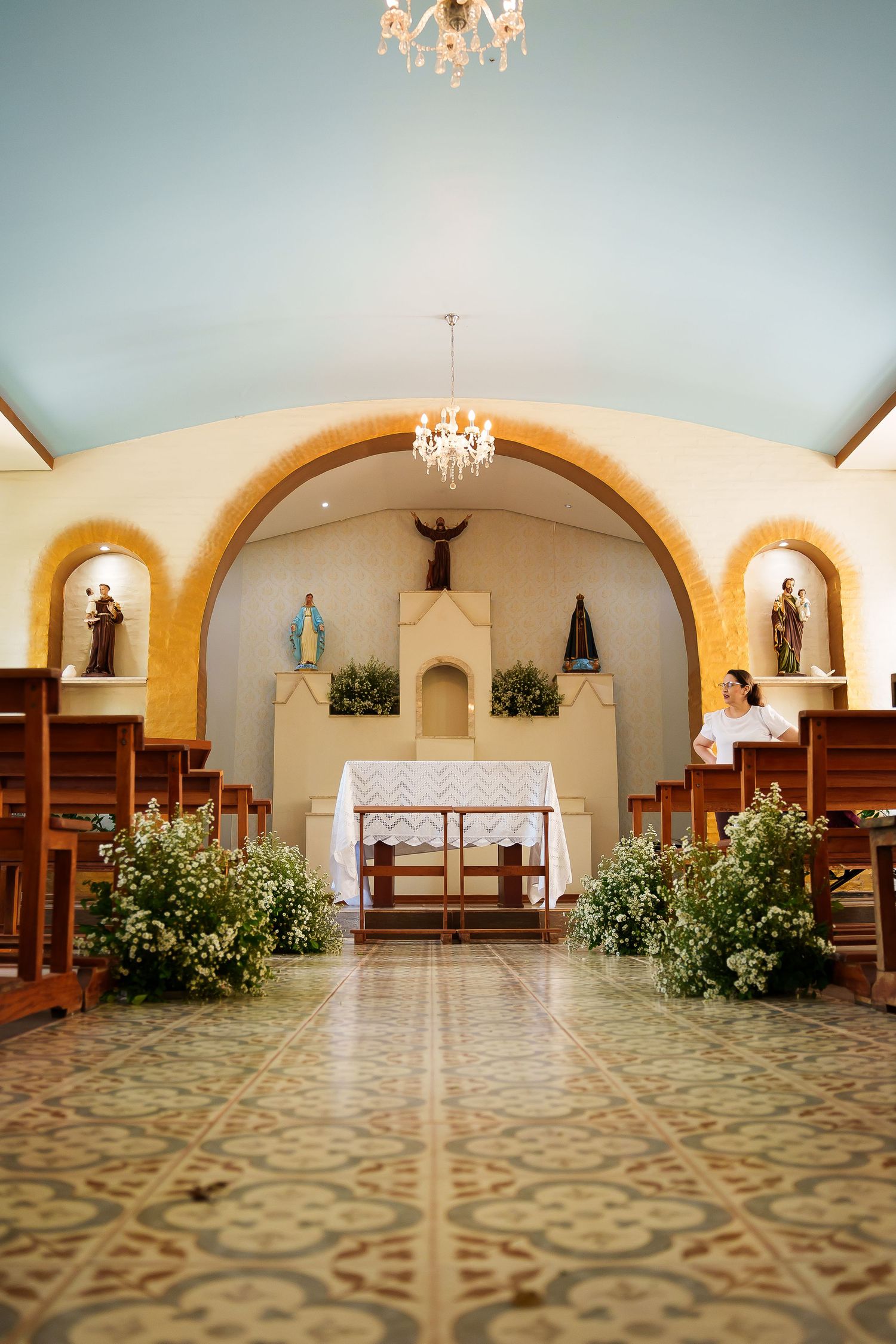 Small chapel interior with wooden pews, floral arrangements, chandelier, and religious statues under arched alcoves.