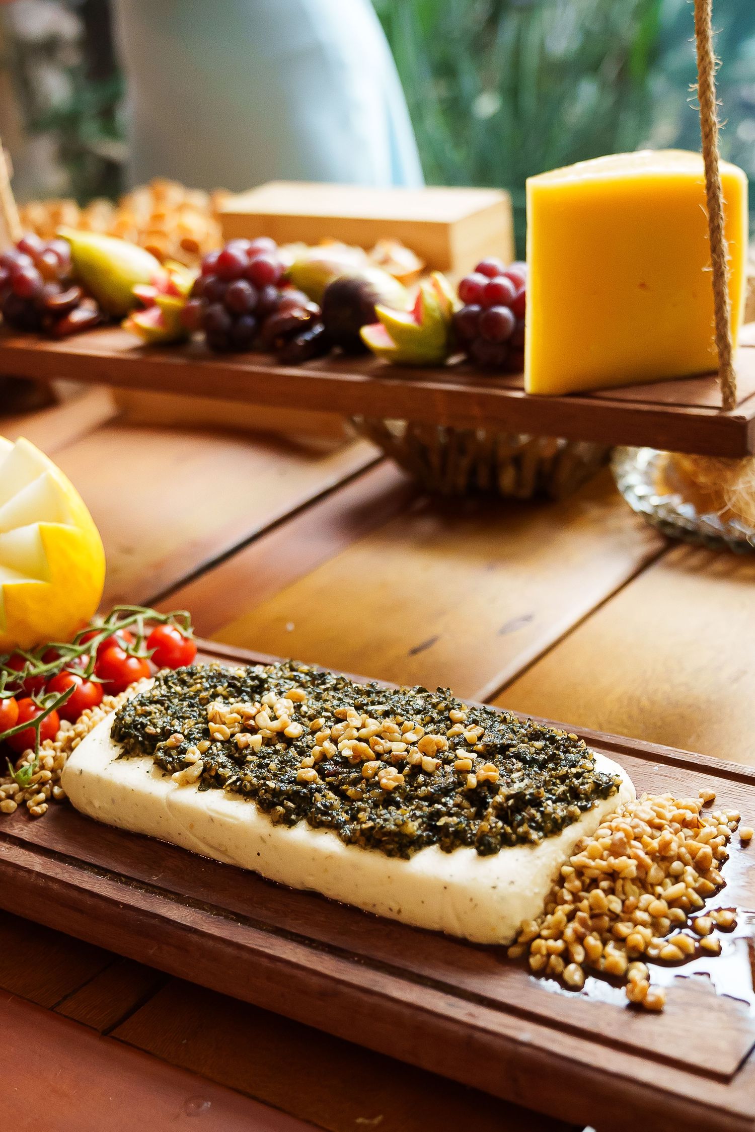 Gourmet cheese board with herb-topped white cheese, yellow cheese wedge, fresh grapes, fruits, and nuts on wooden table.