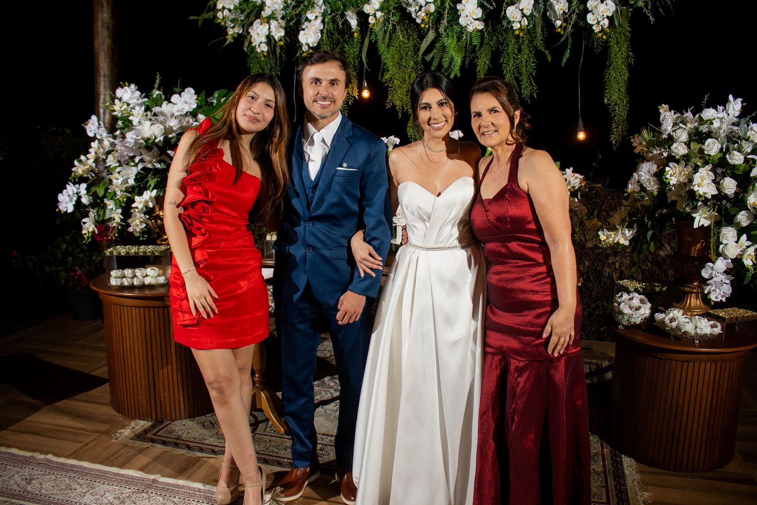 Bride and groom pose with two women in red and burgundy dresses at an elegant outdoor wedding reception.
