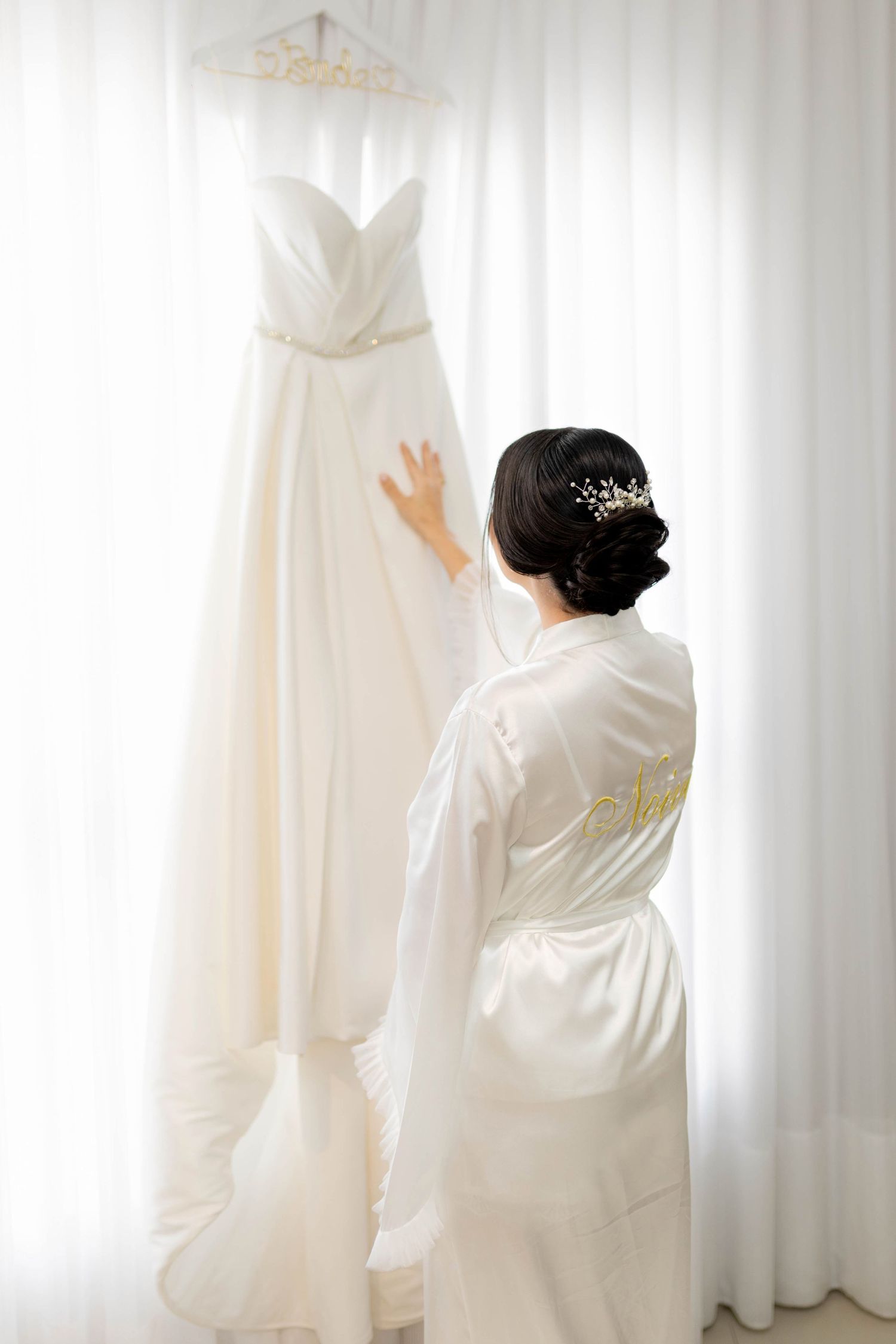 Bride in white satin robe with hair updo admiring her elegant strapless wedding gown hanging by a bright window.