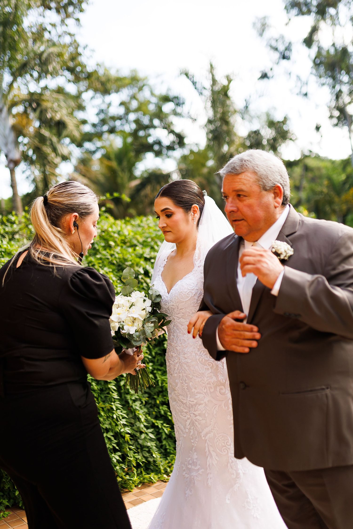 Bride in white lace gown holding bouquet stands with older man in gray suit and woman in black dress outdoors.