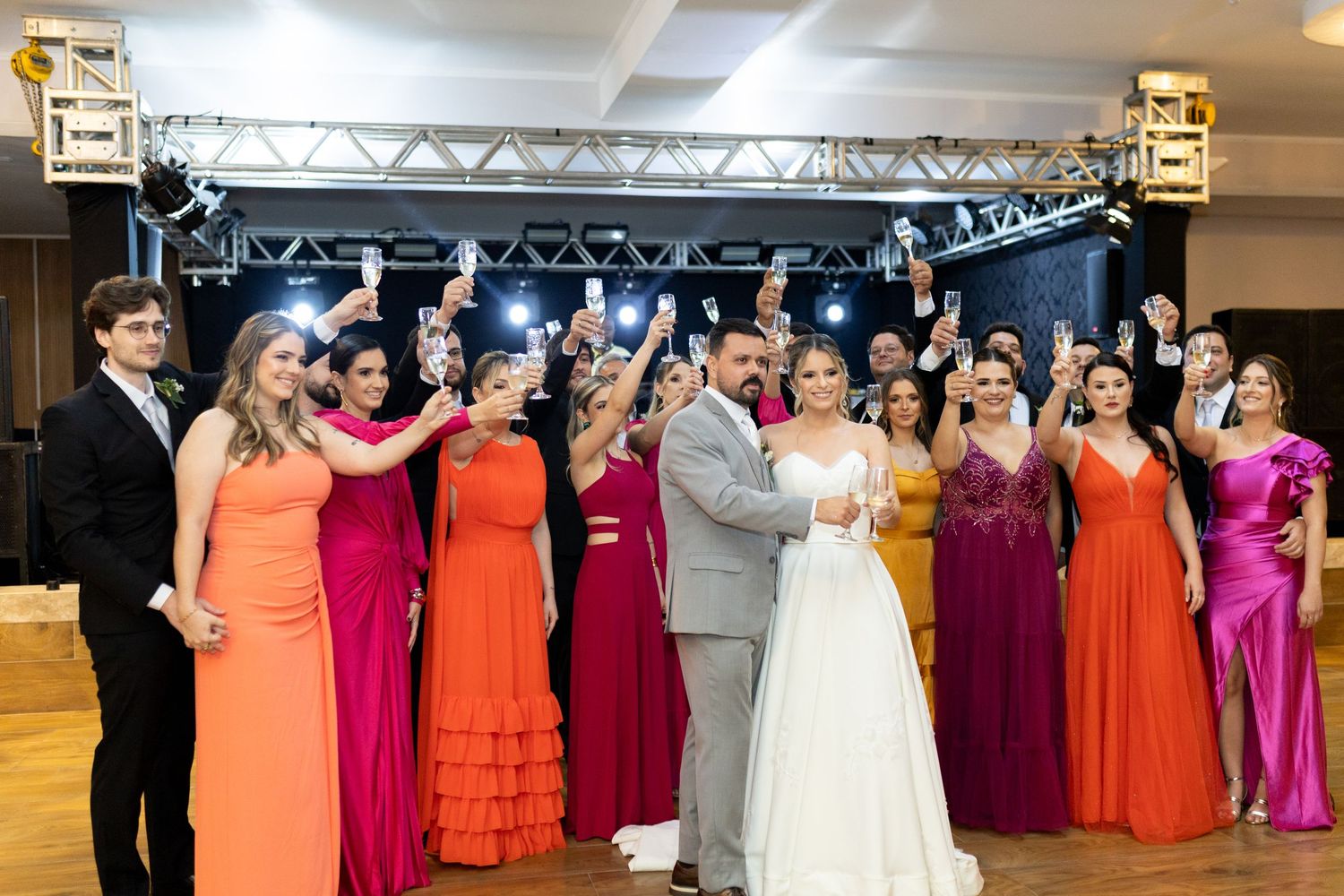 Wedding party celebrating in a ballroom, bride and groom surrounded by guests in colorful red, orange, and pink gowns.