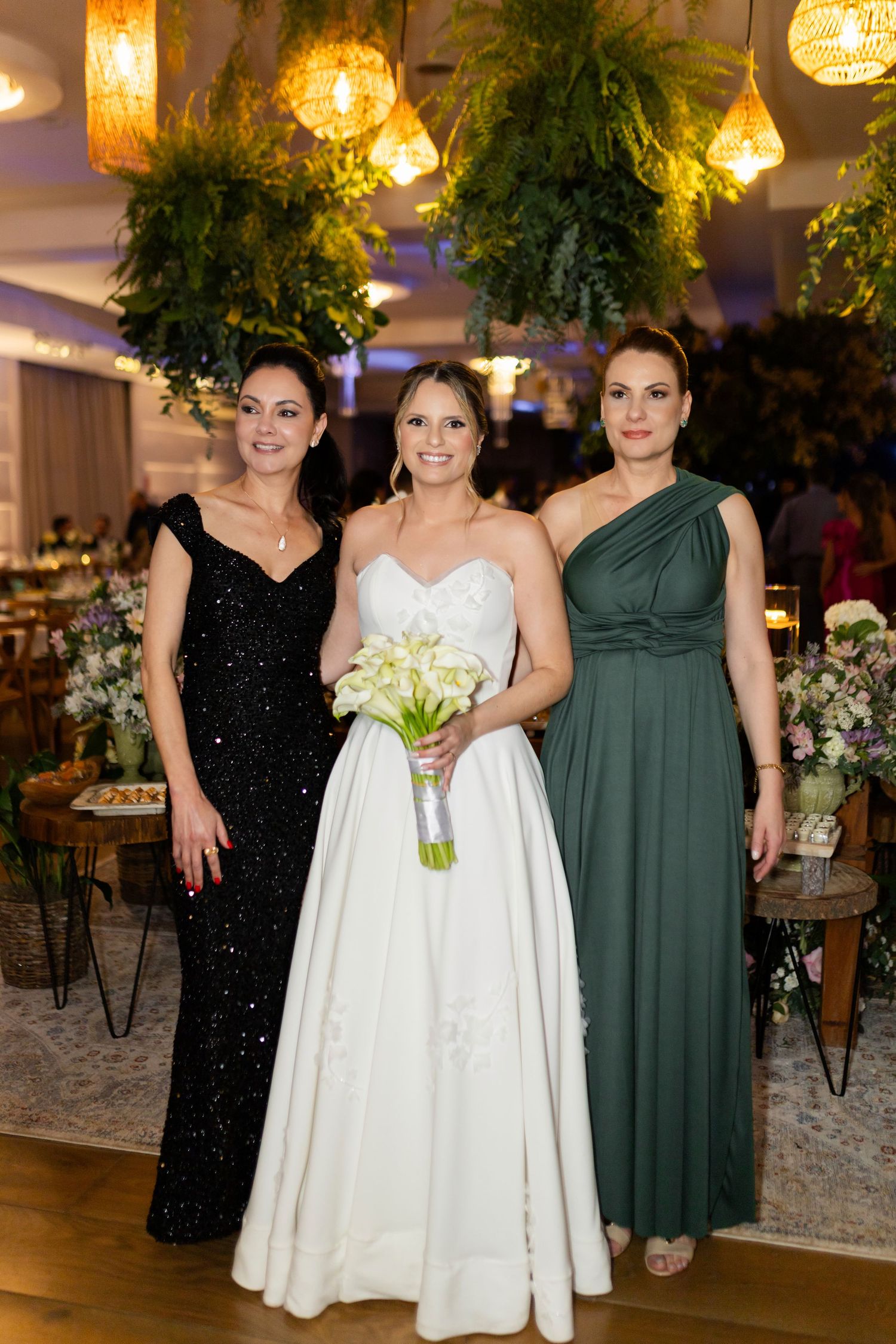 Bride in white gown holding calla lily bouquet poses with two women in black and green formal dresses at elegant wedding reception.