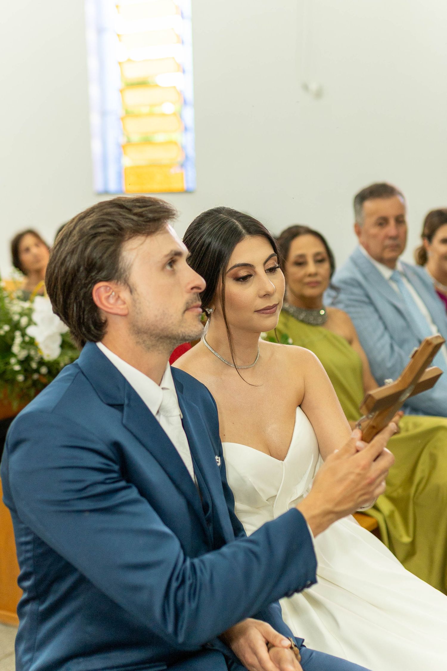 Bride and groom sitting together in church pews during wedding ceremony, groom in blue suit holding wooden cross.