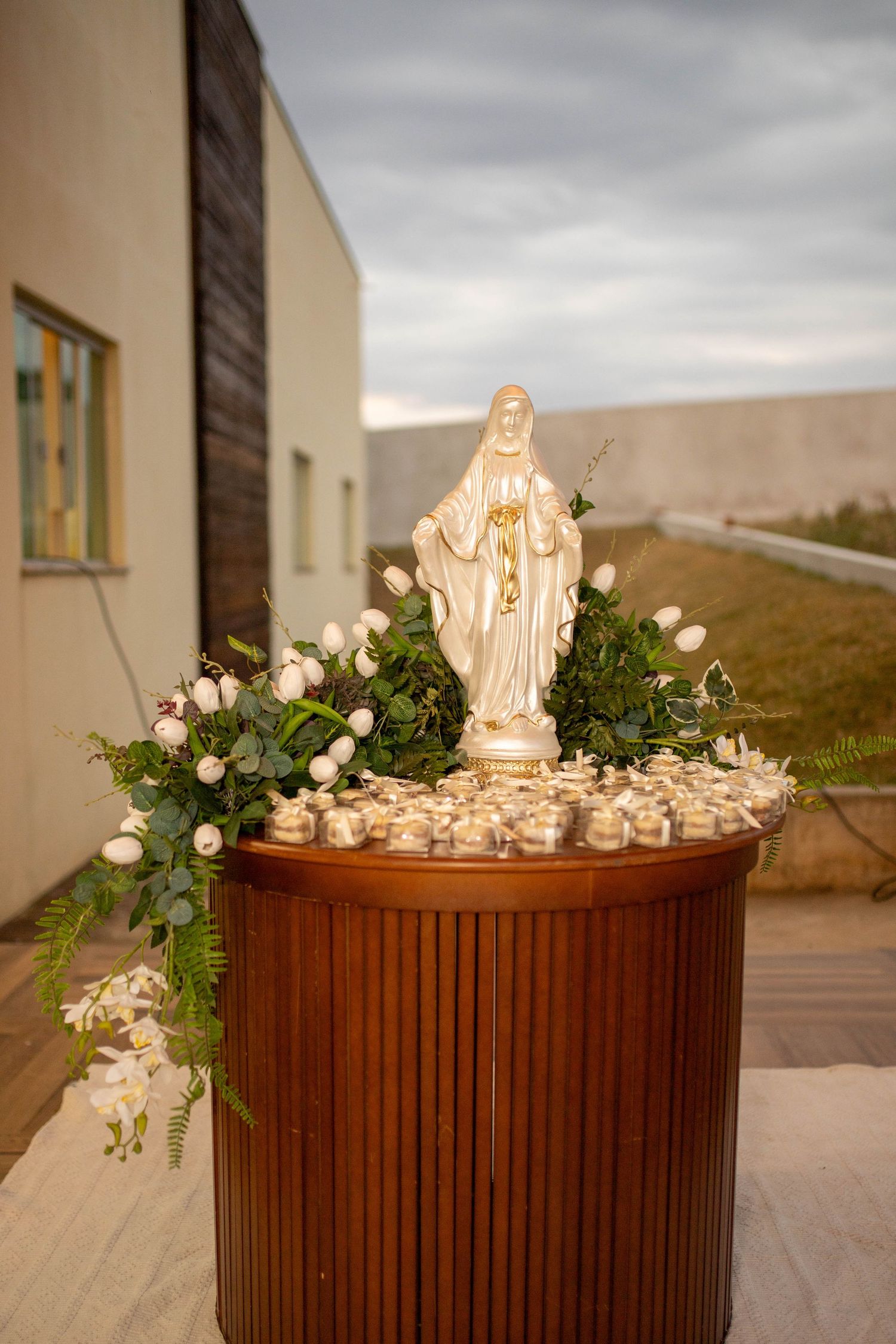 White Virgin Mary statue surrounded by flowers and candles on a wooden pedestal outdoors at a religious ceremony.
