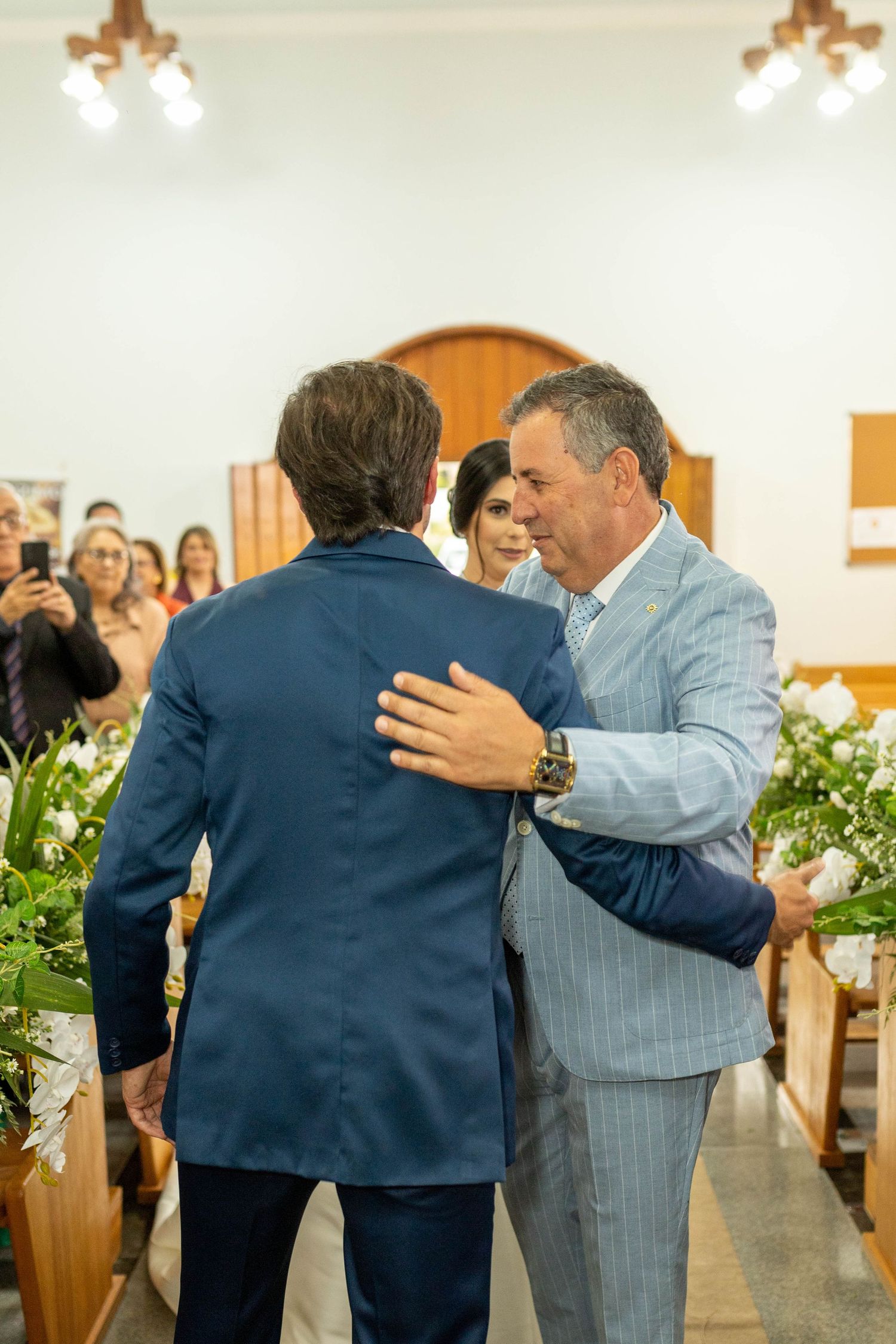 Two men embrace at a church wedding ceremony, surrounded by floral arrangements and wedding guests in the background.