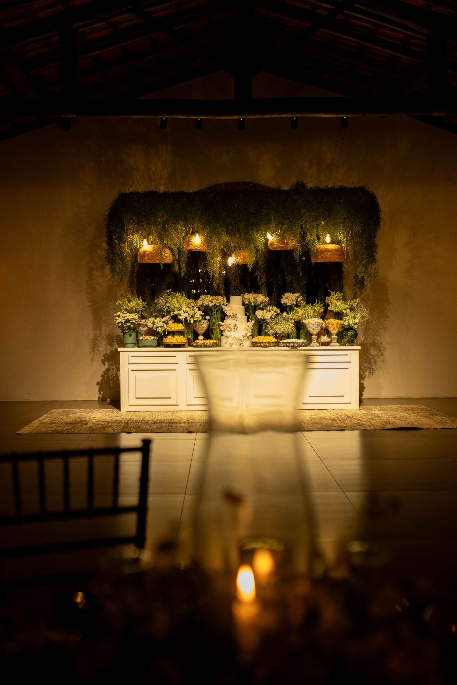 Elegant wedding reception with candlelit dessert table, lush greenery backdrop, and warm ambient lighting in a rustic venue.